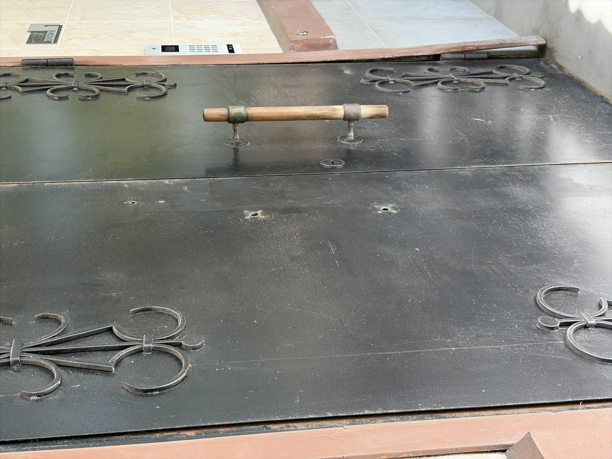 A close-up, eye-level shot shows a large, dark metal double door. The door is divided vertically down the middle, suggesting it is a double door. On the left door panel, a decorative iron scrollwork design is visible towards the top. The right door panel features a wooden handle with metal fittings, positioned horizontally. Below the handle, another, smaller iron scrollwork design is present. Several circular indentations, possibly keyholes or screw holes, are scattered across the door panels. To the right of the doors, a beige tiled wall is visible, featuring a small metal plaque with the number "1" and some text in Cyrillic. Below the plaque, a silver intercom panel with a keypad and screen is mounted on the wall. The bottom of the doors and the wall meet a light gray concrete floor. The scene is outdoors, with what appears to be a building exterior. The lighting is natural and suggests daylight. No individuals are present in the image.