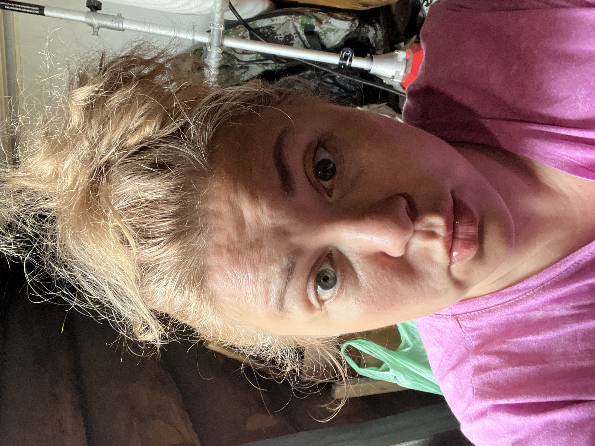 A woman with blonde hair in a messy bun, wearing a pink t-shirt, takes a selfie in what appears to be an attic. Her eyes are wide and her mouth is pursed. Behind her, there is a green plastic bag, a camouflage backpack, and a white pole with a red base. The wooden beams of the attic roof are visible in the background. The overall impression is one of a casual, unposed moment.