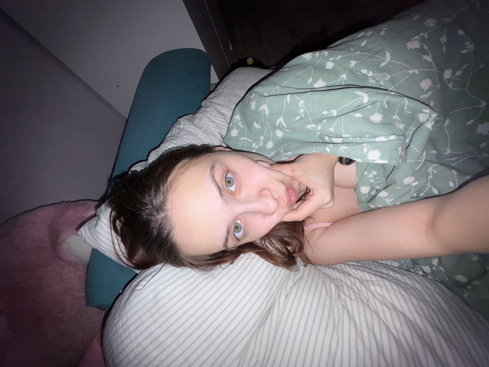 A young woman lies in bed, her head resting on a striped pillow with a blue cushion behind her. She is partially covered by a green floral-patterned blanket. Her right hand is near her face, touching her cheek. The background includes a partially visible pink object to the left and a dark space with a yellow object to the right. The location cannot be confirmed as Прикамский, Russia based on the visual content.