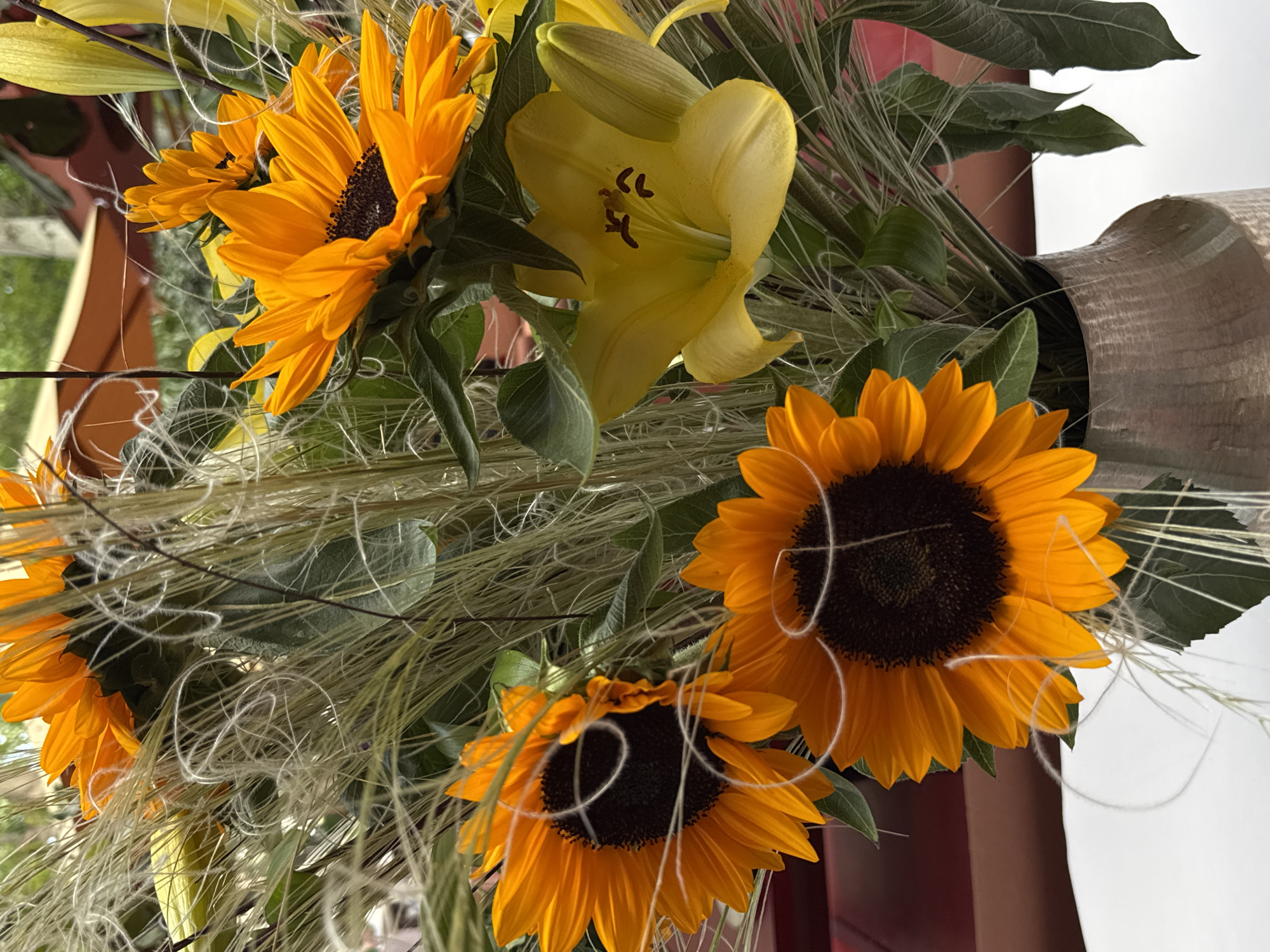 A close-up shot depicts a bouquet of sunflowers and a yellow lily in a vase. The scene is well-lit, highlighting the textures and colors of the flowers. The sunflowers have large, dark brown centers surrounded by bright orange petals. The yellow lily is partially open, showing its inner details. The bouquet includes leafy green foliage and decorative wispy strands. The vase is made of a light brown, textured material. The background is a mix of white and red, suggesting an interior setting. The location cannot be determined from the image.