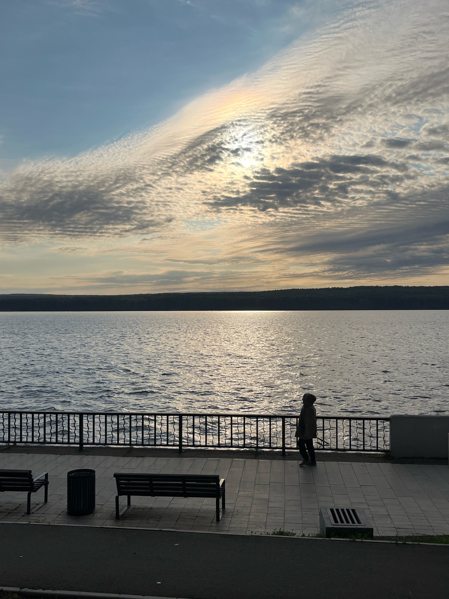 The image depicts a serene waterside scene at what appears to be a public promenade in Chaikovsky, Russia. The foreground features a paved walkway with two dark-colored park benches and a cylindrical trash receptacle. A metal railing borders the promenade, separating it from a large body of water. In the middle ground, the water stretches out, reflecting the sky and a distant tree-lined horizon. The sky above is a canvas of soft blues and grays, with dramatic, striated clouds catching the light of the setting sun. A faint iridescence, possibly a sun dog, is visible on the right side of the sky. On the promenade, a lone figure, silhouetted against the water and sky, stands facing the lake, appearing to be contemplating the view or perhaps fishing with a rod. The overall atmosphere is tranquil and contemplative.