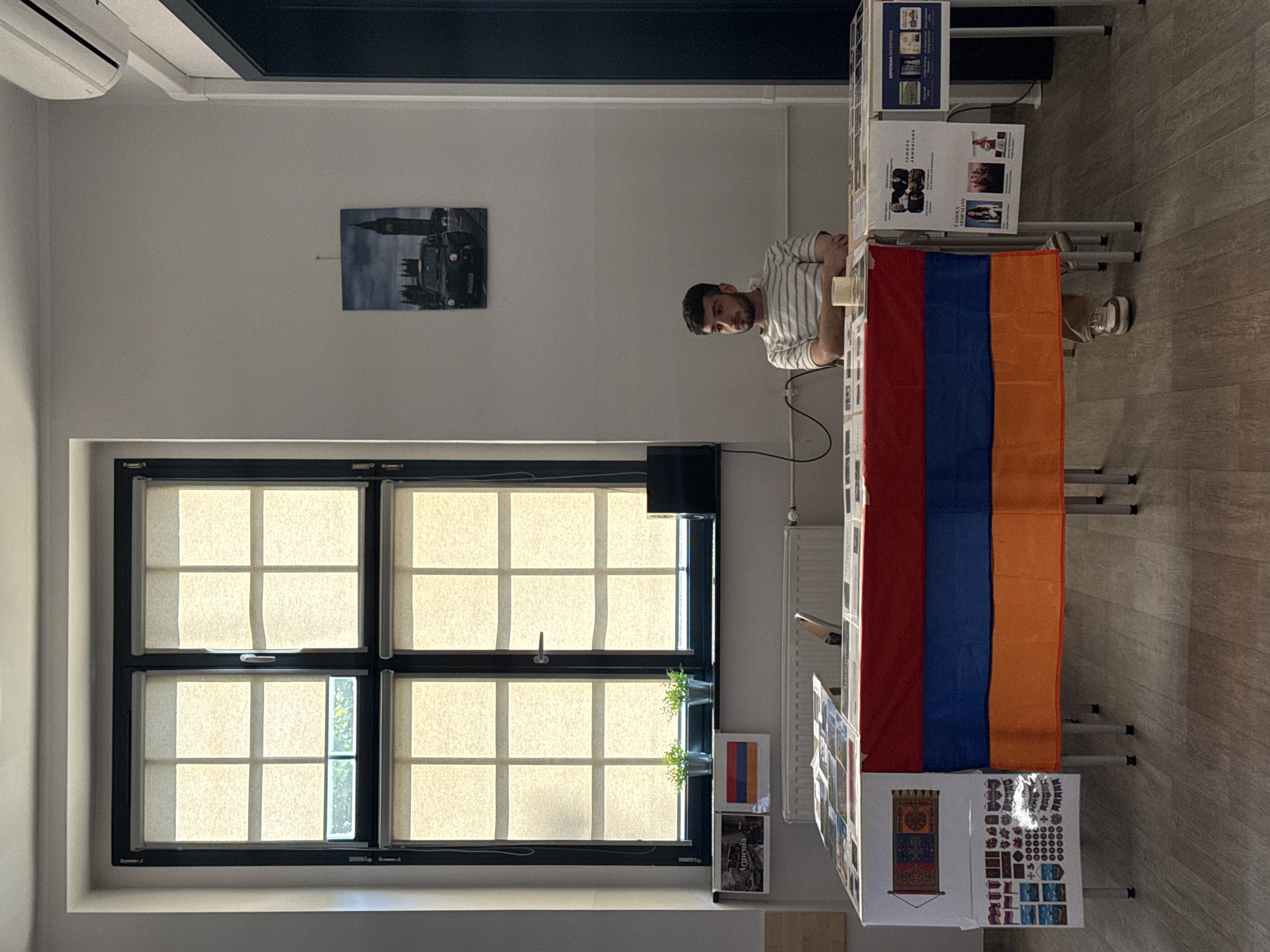 An indoor scene at what appears to be a cultural event or exhibition in Opole, Poland. A young man with dark hair and a beard, wearing a striped t-shirt and beige pants, sits behind a table. He is looking to his left, with his arms crossed. The table is draped with an Armenian flag and displays various items, including what look like printed photographs, posters, and small flags. The backdrop features a large window with blinds, a radiator, and a framed picture of a London taxi and Big Ben on the wall. The lighting suggests an overcast day or an indoor setting with artificial lights.