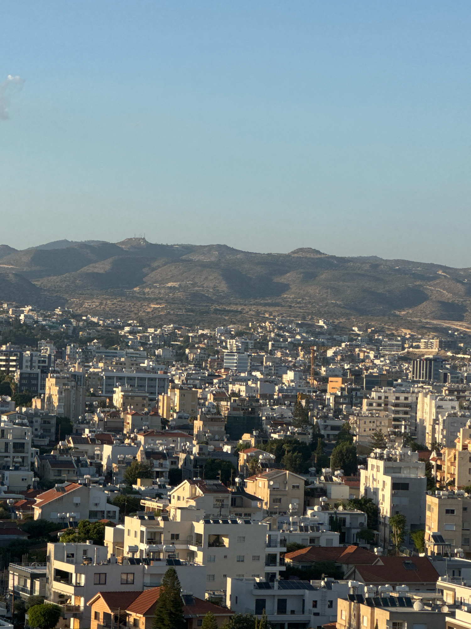 The image shows a panoramic view of Limassol, Cyprus, a coastal city characterized by its densely packed residential and commercial buildings. In the foreground, a cluster of white and beige buildings with red-tiled roofs dominates the scene. These structures, varying in height, display architectural features typical of Mediterranean regions, including balconies and prominent solar water heaters on their rooftops.
Further into the cityscape, taller buildings emerge, including modern apartment blocks and office structures, some with glass facades reflecting the sunlight. A prominent orange construction crane is visible amidst the buildings, suggesting ongoing urban development. The urban sprawl extends towards a range of rugged, arid hills that form the backdrop of the city. These hills are sparsely vegetated with dry shrubs and grasses. A prominent structure, possibly a telecommunications tower or a small settlement, sits atop one of the highest peaks in the distance. The sky above is clear and a pale blue, indicating it is likely daytime, with the warm glow of the sun suggesting either early morning or late afternoon. The overall impression is of a bustling city integrated with its natural landscape, bathed in the soft light of the Mediterranean sun.