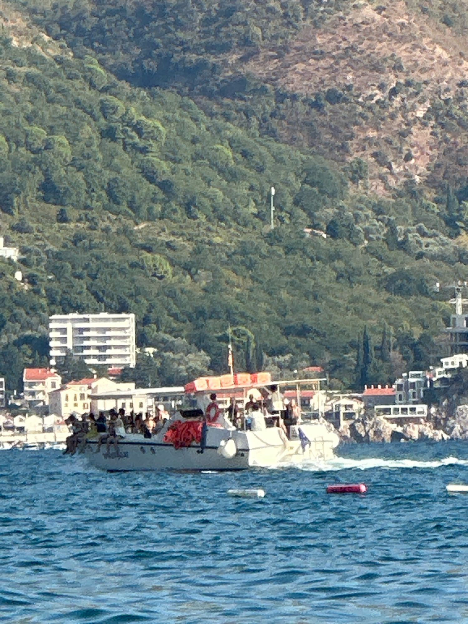 The image captures a daytime scene at a coastal location, likely in Boreti, Montenegro, given the context. In the foreground, the choppy blue water is visible, with several floating objects, including a pink inflatable raft. A white boat, carrying approximately 15-20 people, is the central focus in the mid-ground. The passengers, some appearing to be tourists, are seated along the sides of the boat, with some standing. The boat is moving, creating a wake.
Behind the boat, a cluster of buildings forms the coastline. A prominent white, multi-story building with numerous horizontal windows is situated on a gentle slope. Several other buildings with red-tiled roofs are also visible, nestled amongst trees. In the background, a densely vegetated hillside rises steeply, covered in a variety of green foliage. The overall impression is a bustling waterfront area with leisure activities taking place.