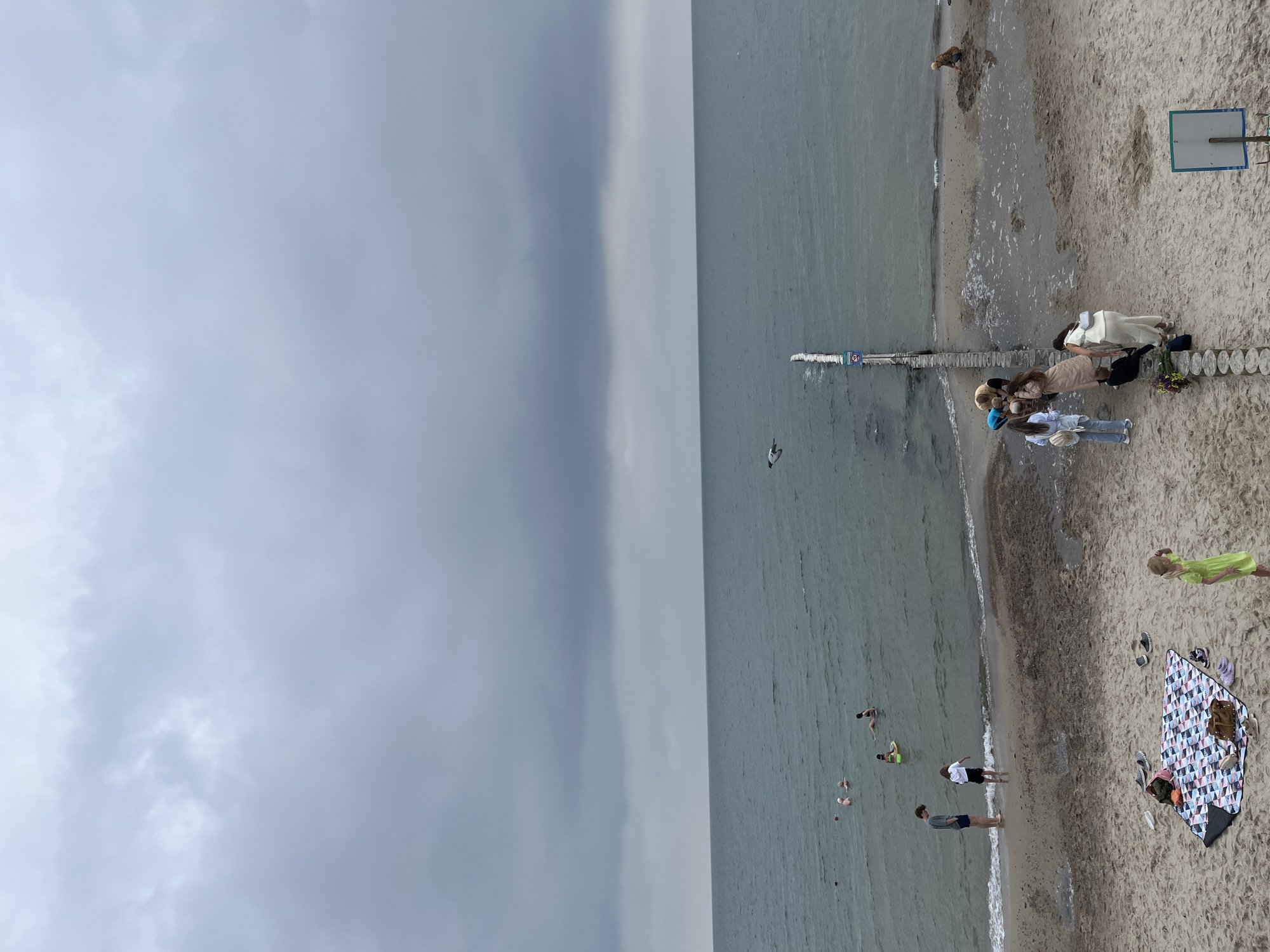 The image shows a cloudy day at a beach in Zelenogradsk, Russia. In the foreground, on the sandy beach, a picnic blanket with various items is spread out. To the right, a young girl in a lime green dress stands with her hand raised to her head. Further back, a group of people are gathered near a wooden pier that extends into the sea. Several individuals are wading or swimming in the water, with some on flotation devices. In the distance, the sea meets the horizon under a heavy, overcast sky. A single bird is visible in flight over the water.