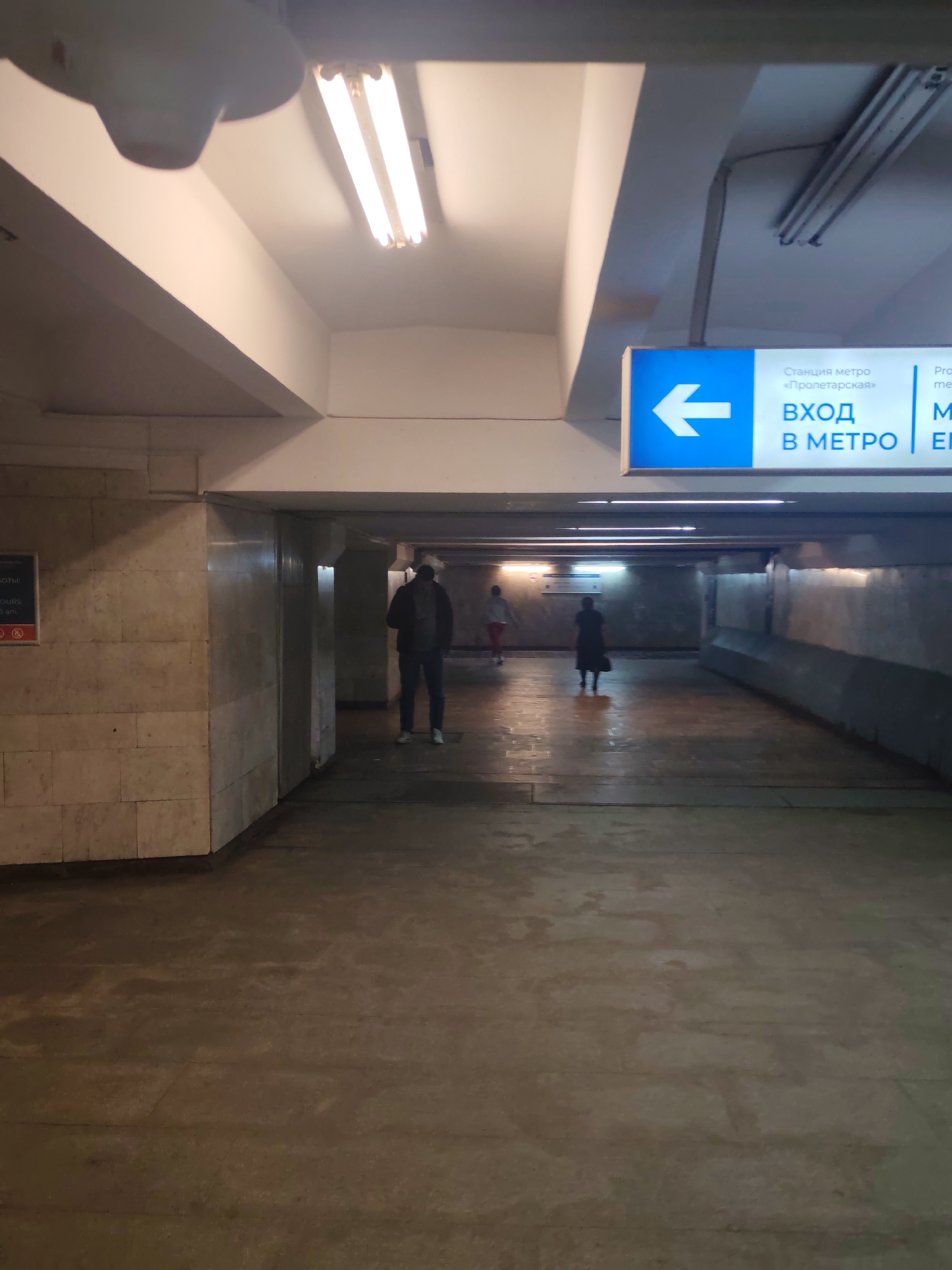 An underpass in Nizhny Novgorod, Russia, features a sign with a blue background and white text pointing left, indicating "Entrance to the Metro." The underpass has a concrete floor and walls, with overhead fluorescent lighting. Several people are present, including a man walking towards the camera, and others further down the underpass. The overall atmosphere is dimly lit and somewhat utilitarian.