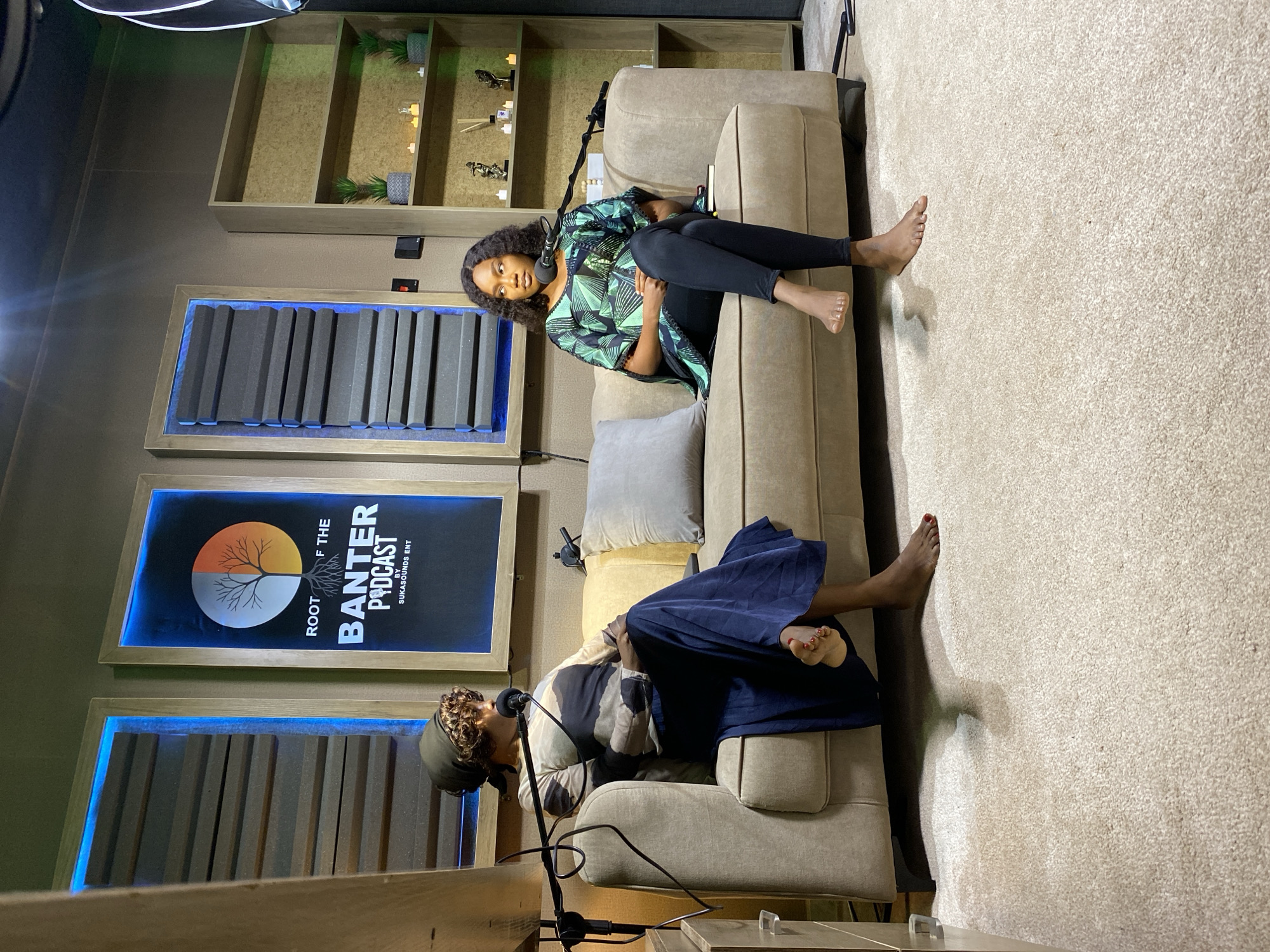 Two women are sitting on a plush, light brown sofa in a room. The room appears to be a podcast studio. The walls are a muted brown, with wooden paneling. Behind the sofa, there are two large framed panels with acoustic foam, one of which is illuminated with blue light. To the right of these panels, there is a bookshelf with a few potted plants and decorative items.
The woman on the left is wearing a long, dark blue skirt and a long-sleeved shirt with a patterned design in shades of brown and cream. She has a headscarf tied around her head. She is leaning back on the sofa, with her left arm resting on the backrest and her right arm on her lap. A microphone is positioned in front of her. Her bare feet are visible, with red nail polish on her toes.
The woman on the right is wearing black leggings and a short, patterned green and black top. Her hands are clasped in her lap, and she is looking forward. A microphone is also positioned in front of her. Her bare feet are visible, crossed at the ankles.
The floor is covered in a thick, cream-colored carpet. There are two light-colored throw pillows on the sofa. The lighting in the room is dim, with a spotlight shining from above, creating some shadows. The overall atmosphere suggests an indoor recording session. The location is not specified within the image or its accompanying text.