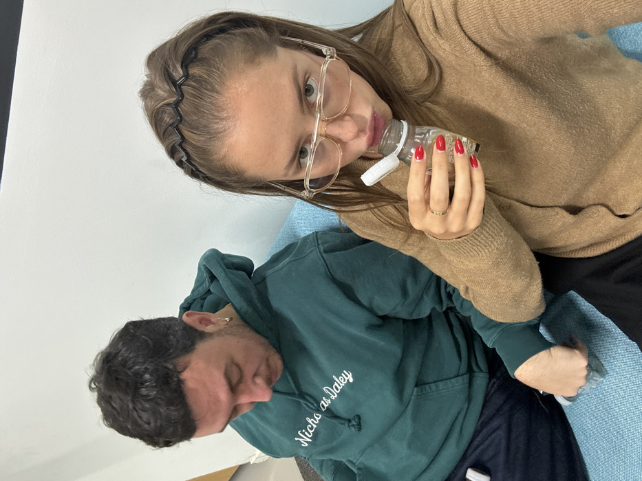 Two individuals are seated indoors on a light blue upholstered bench against a plain white wall. In the foreground, a female with light brown hair, wearing a black zigzag headband and clear-framed glasses, is dressed in a light brown cardigan. Her right hand, adorned with red nail polish and a gold ring, holds a small, clear plastic squeeze bottle containing a yellowish liquid, from which she is actively drinking. Her gaze is directed towards the camera. To her left and slightly behind, a male with dark curly hair, light facial stubble, and a gold hoop earring in his left ear wears a dark teal hooded sweatshirt with "Nicholas Daley" written in white script. He looks downwards and to his right, holding a small, light-colored object in his right hand. The "Nicholas Daley" branding on the male's apparel indicates the scene is likely located in London, United Kingdom.