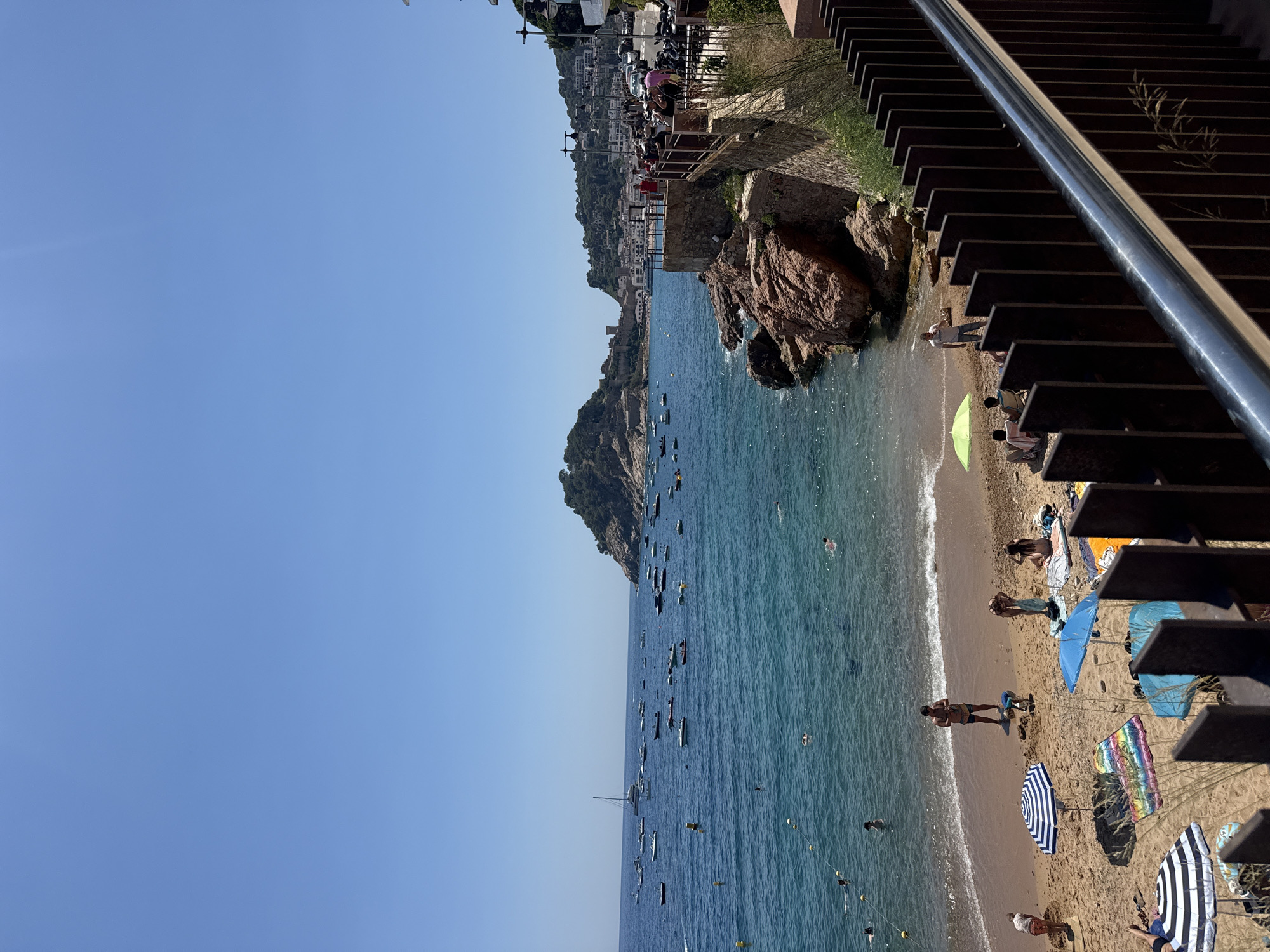 This image depicts a sun-drenched scene at a beach in Tossa de Mar, Spain. The azure water is dotted with numerous boats of varying sizes, from small dinghies to a larger sailboat. Several people are visible swimming and wading in the clear, shallow water near the shoreline.
The sandy beach is occupied by a small number of beachgoers. Some are sunbathing under colorful umbrellas, while others are interacting with each other or engaged in activities like building sandcastles. Beach towels and other personal items are scattered across the sand.
To the right of the beach, a rocky outcropping juts into the sea, topped with a stone wall and a walkway lined with a metal railing. People are seen strolling along this walkway, observing the beach and the water. Beyond the walkway, the town of Tossa de Mar unfolds, with buildings climbing the hillside and a prominent castle-like structure visible on a distant hill. The overall impression is one of a vibrant coastal resort on a clear, bright day.