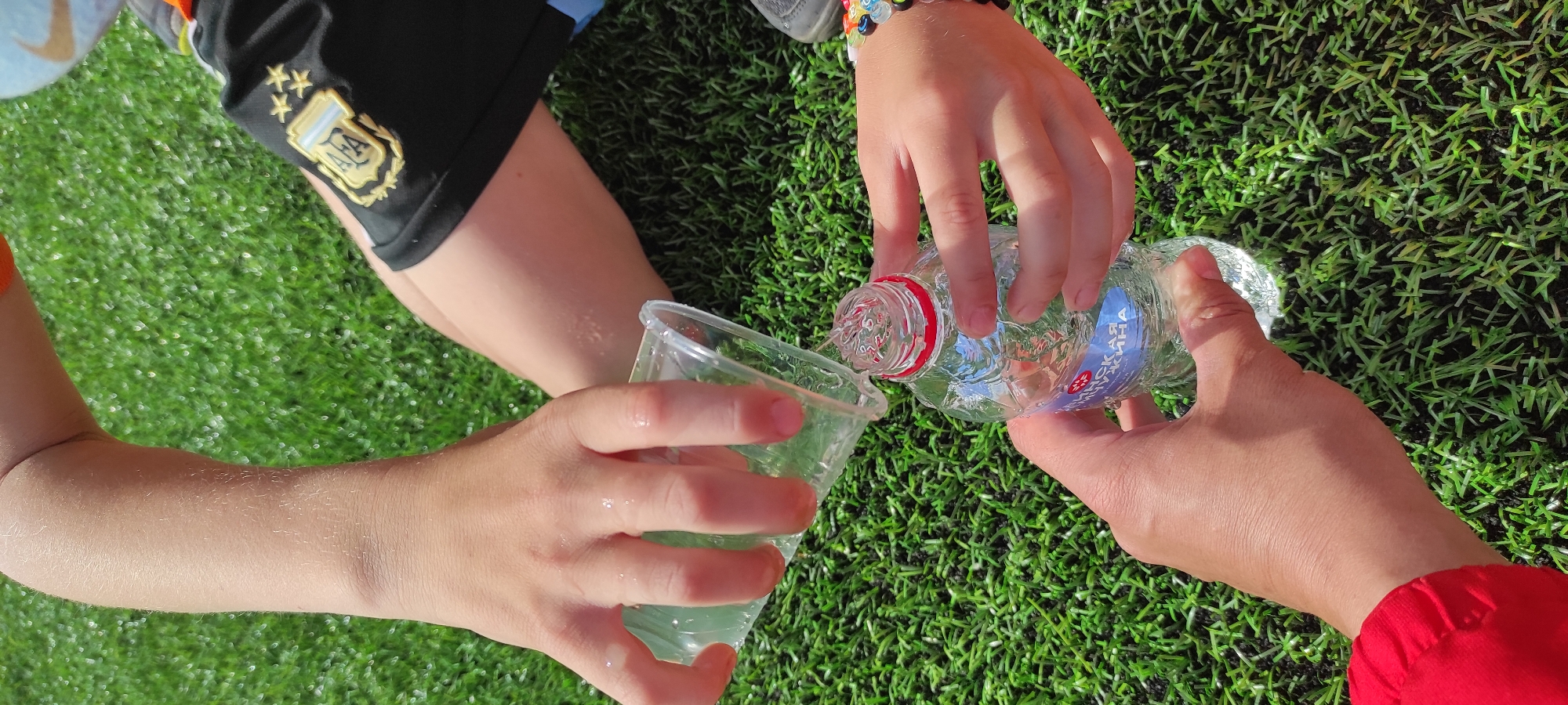 In Noviy, Russia, on a bright, sunny day, an outdoor scene unfolds on a vibrant green, artificial turf field. Two sets of hands are engaged in the action of pouring water. The primary focus is on the hands of two individuals, with a third hand visible at the bottom of the frame. One person's hands are holding a small, clear plastic cup, from which clear liquid is being poured into the opening of a clear plastic water bottle. The bottle has a red cap and a blue and white label. The second person's hands are gripping the water bottle to steady it. The person on the right wears a black sports jersey with a prominent crest and the number "10" visible, along with colorful beaded bracelets on their wrist. The person on the left wears a bright red garment, possibly a jacket. The background is entirely the green turf, suggesting a sports field or park. The overall impression is a moment of hydration or a playful activity taking place outdoors.