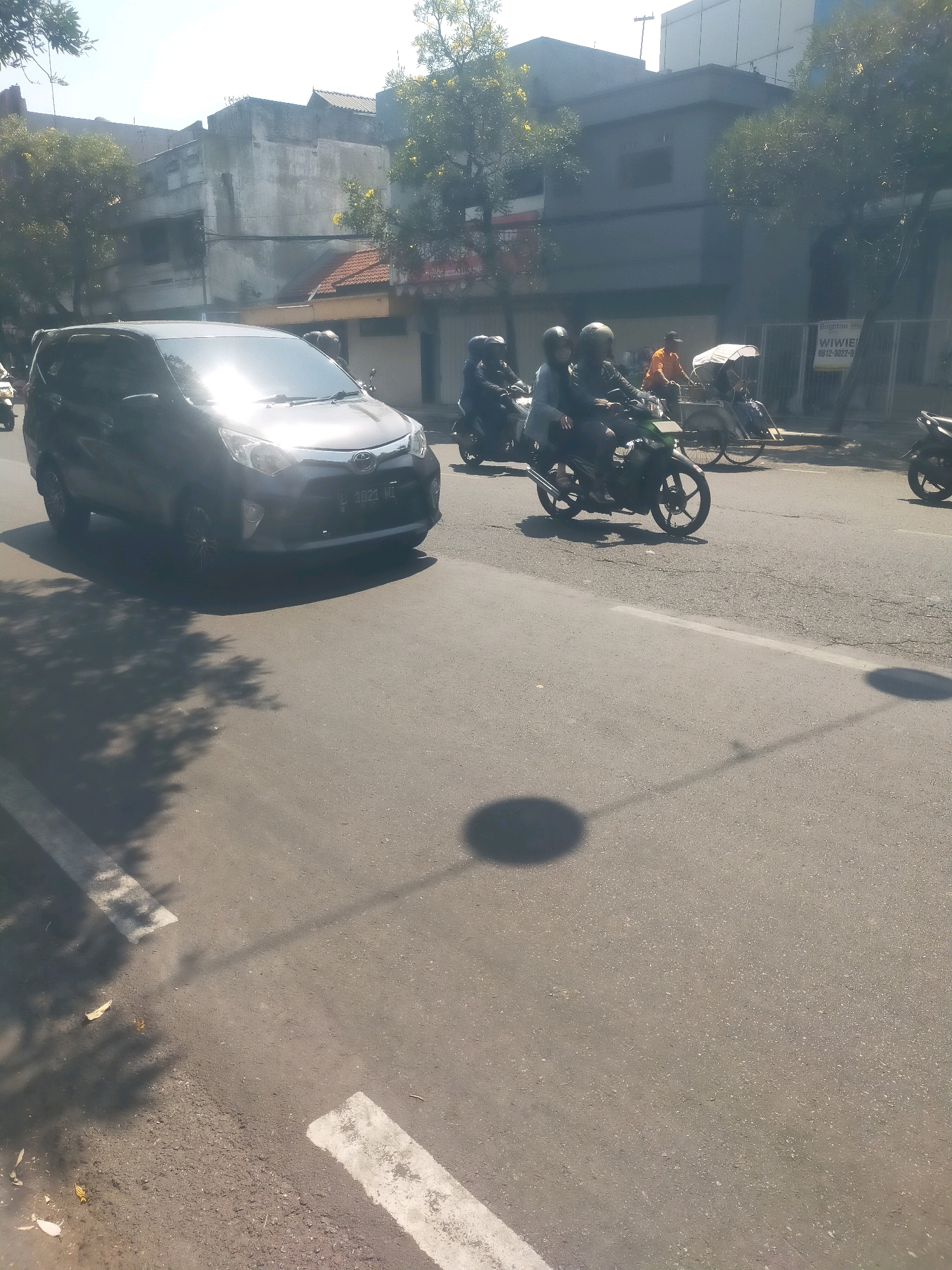 In Sidoarjo, Indonesia, a dark gray car drives on an asphalt road under a bright sky. The car is positioned on the left side of the frame, facing right. Behind the car, two motorcycles with riders, each wearing helmets, travel in the same direction. One motorcycle has a driver and passenger, while the other appears to have only a driver. Further down the road, a man in an orange shirt drives a pedicab. In the background, buildings and trees line the street. A sign reading "BRIGHTON" is visible on a fence. Shadows from trees and a circular object, possibly a lamppost, are cast on the road.