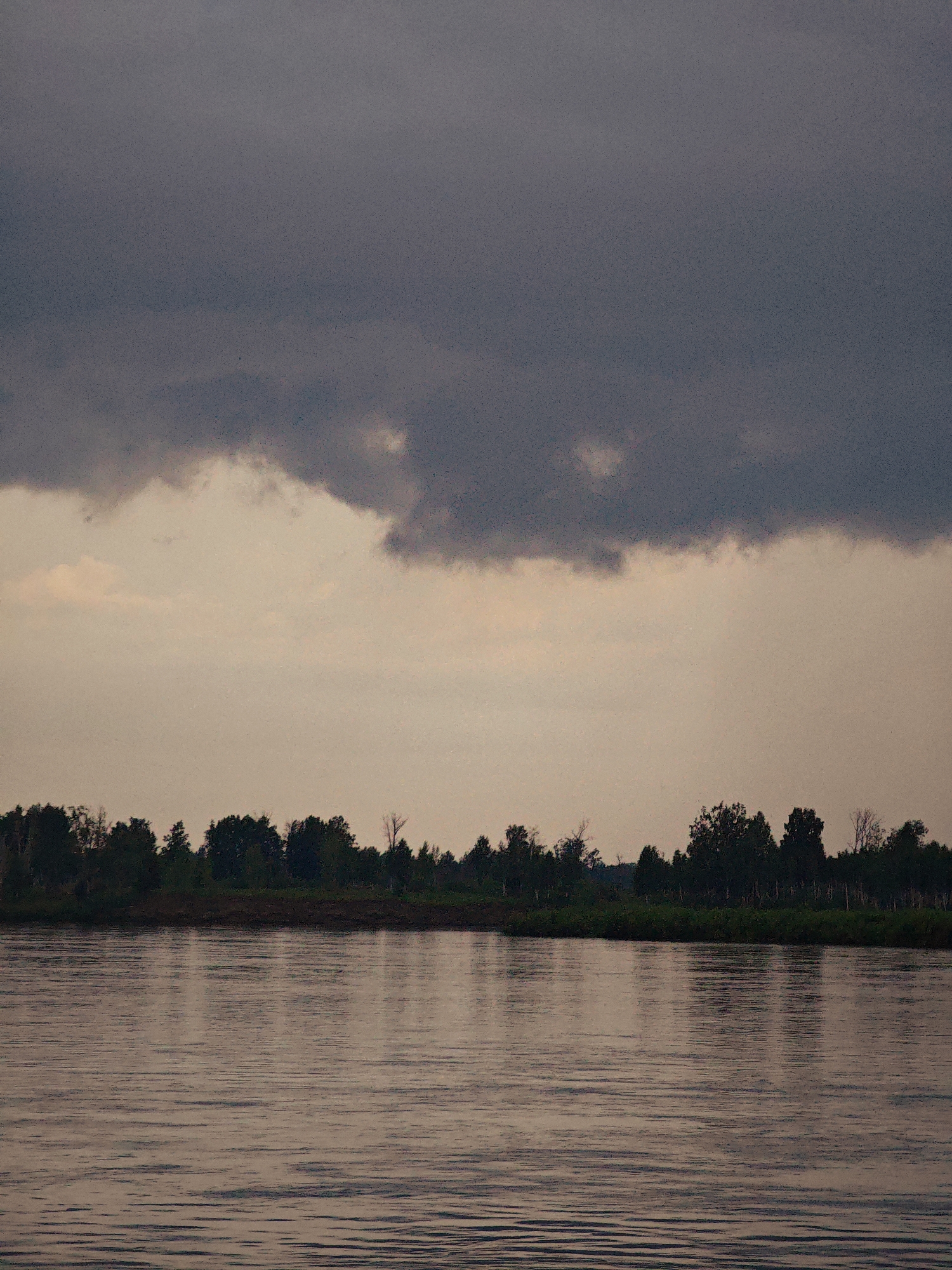 The image shows a wide, calm river with gentle ripples reflecting the subdued light of the sky. The sky is dominated by a large, dark cloud bank in the upper portion, with lighter, patchy clouds below it. Along the far bank of the river, a dense line of dark green trees forms a silhouette against the pale sky. The scene appears to be natural and undeveloped, with no signs of human structures or activity. The location is identified as being in the vicinity of the city of Kazachinskoye, Russia, likely a rural or natural area adjacent to the Yenisei River.