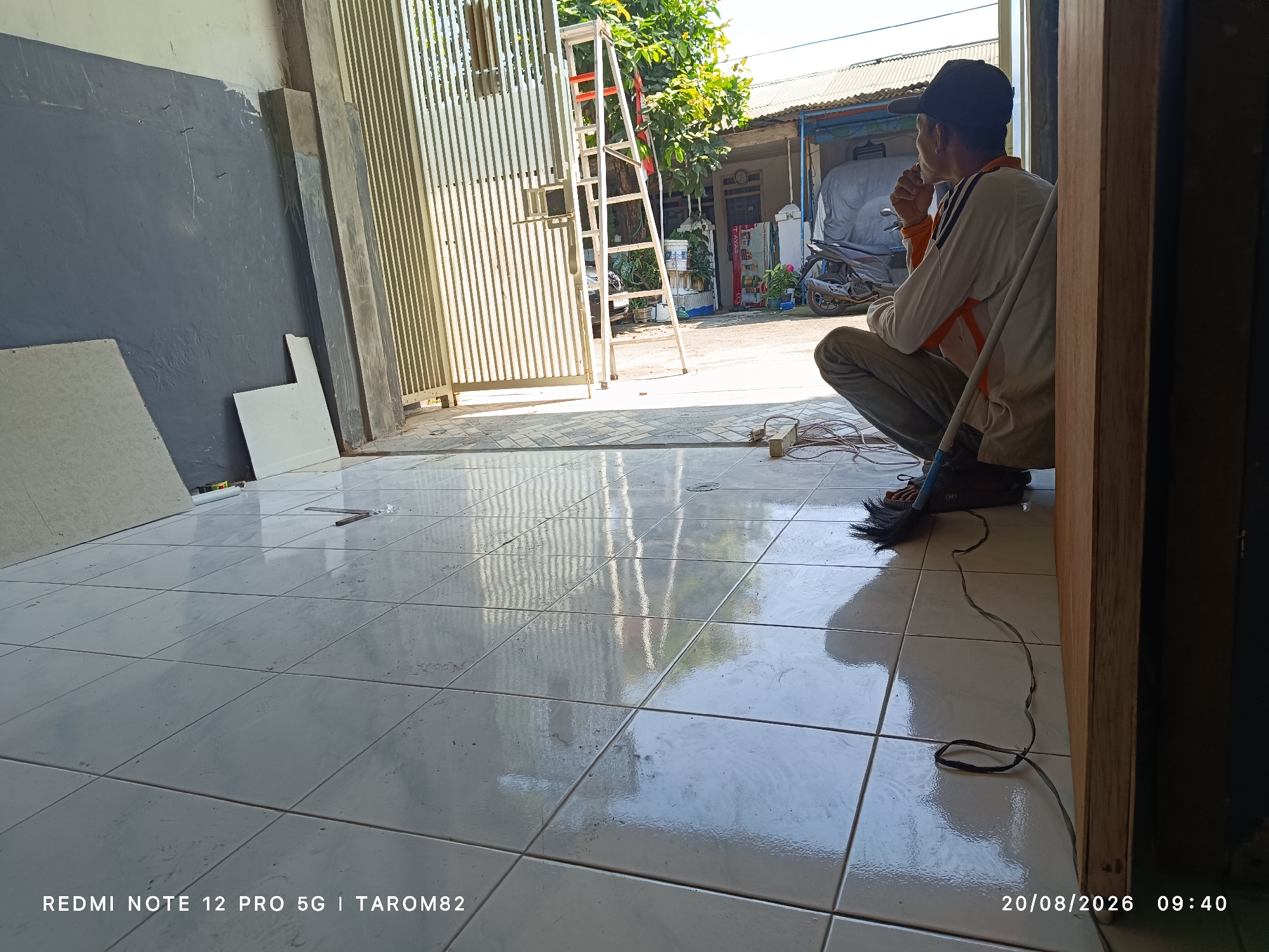 The image depicts an outdoor scene in Sruni, Indonesia, with a focus on a man sitting on the ground. The man, wearing a black cap, an orange and white long-sleeved shirt, and grey pants, is seen from the side in a squatting position. He appears to be contemplating, with his hand near his chin. A white pole is beside him, and electrical cords are on the tiled floor.
The foreground consists of light-colored, reflective square tiles, showing reflections of the surrounding environment, including a metal gate and foliage. Behind the man, a ladder leans against the metal gate. The background reveals a residential area with houses and a motorcycle parked near a building covered with a tarp. The overall setting suggests a construction or maintenance area due to the presence of the ladder and materials. The timestamp "20/08/2026 09:40" is visible in the bottom right corner.
