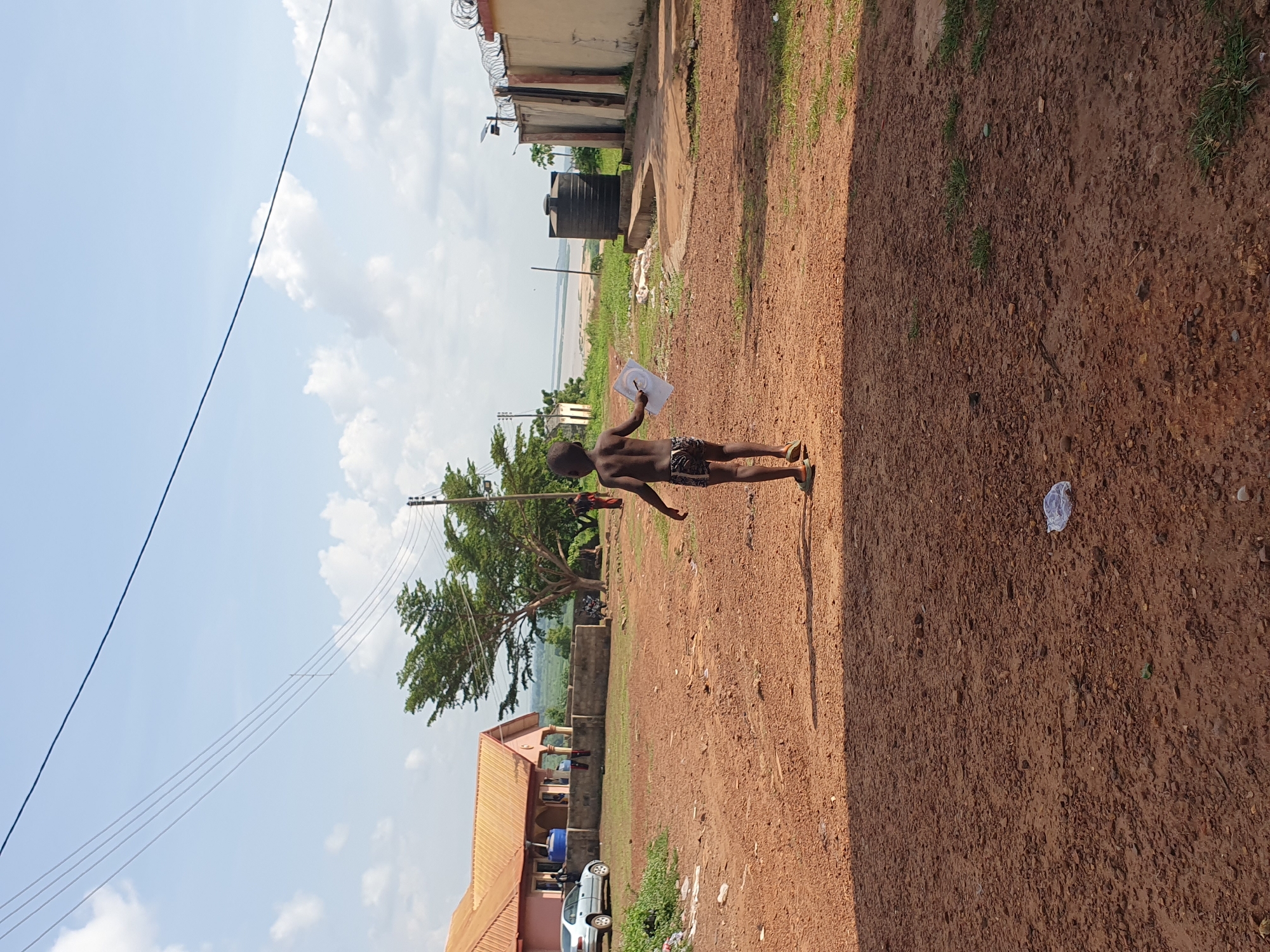 The image depicts an outdoor scene in what appears to be a semi-urban or rural setting in Idah, Nigeria, under bright daylight. The ground is an unpaved, reddish-brown dirt path with scattered patches of green grass. Shadows are visible on the ground, indicating a sunny day.
In the foreground, a young boy is walking or standing on the dirt path. He is shirtless, wearing patterned shorts and flip-flops, and holds a white, thin object, possibly paper or plastic, which has some faint, dark markings or a drawing on it. His back is mostly towards the viewer, and he faces towards the left side of the frame (if rotated correctly).
In the midground, several structures are visible: a building with a terracotta-colored roof and light-colored walls, near which a silver car is parked. Further back, another simpler building has a large, black water tank on its roof, and its perimeter wall is topped with barbed wire. A large, leafy green tree stands prominently, and utility poles with numerous power lines stretch across the sky.
In the distance, beyond the buildings and the tree, a wide body of water, likely a river (consistent with Idah's location on the Benue River), is visible under a hazy sky. The sky above is bright blue with scattered white clouds. The overall scene suggests a typical daily moment in the region. There is no clearly legible text visible in the image.