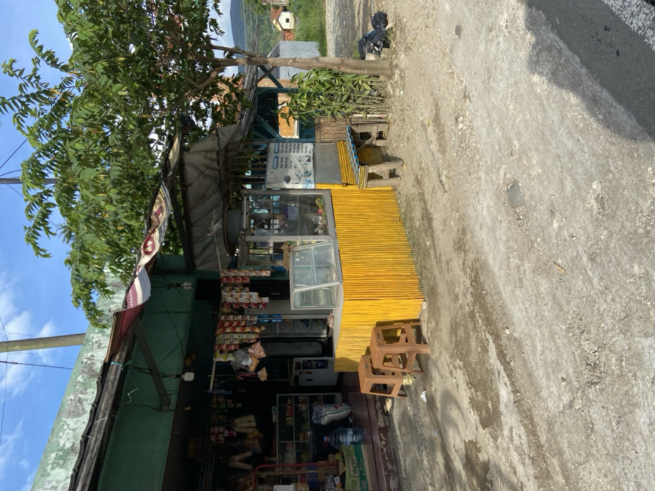 The image shows a small storefront in Sukatani, Indonesia, located at coordinates -6.639 latitude and 107.408 longitude. The storefront is made of wood and has a yellow bamboo fence. There are two brown plastic stools in front of the storefront. There is a glass display case inside the storefront and an open awning above it. There is a tree to the right of the storefront, which is a large tree with green leaves. There are also two small trees in the background, and there is a large green area to the right, which appears to be a park. There is a black bag in the grass to the right. The ground in front of the storefront and to the right is made of gravel and dirt. The sky is blue with white clouds. The front of the storefront appears to be a small food stall. The stall has signs with prices and items, which are in Indonesian. The storefront is closed. There are no people present in the image.