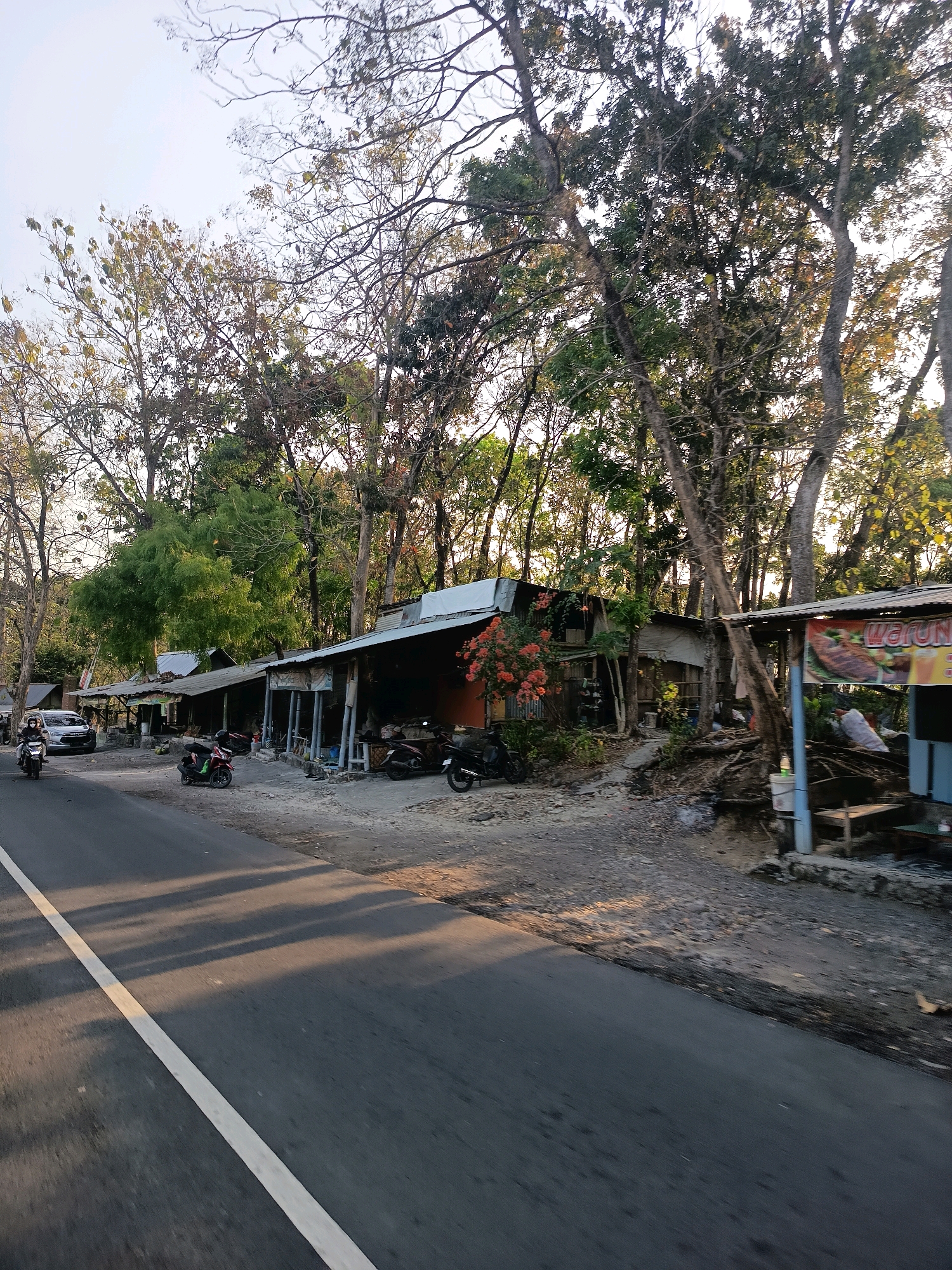 A roadside scene in Ngargosari, Indonesia, features a row of small, rustic shops with corrugated metal roofs, typical of a rural or suburban area. The shops are situated along a paved road, with a white line marking the lane. A silver car is parked on the left side of the road, with a person on a motorcycle driving past it. Several other motorcycles are parked in front of the shops. Lush trees with sparse foliage line the background, some showing signs of seasonal dryness, suggesting a tropical climate. Sunlight casts long shadows across the road, indicating late afternoon or early morning. One shop has a banner with food imagery, implying a food vendor. The overall atmosphere is one of a quiet, local commercial area.