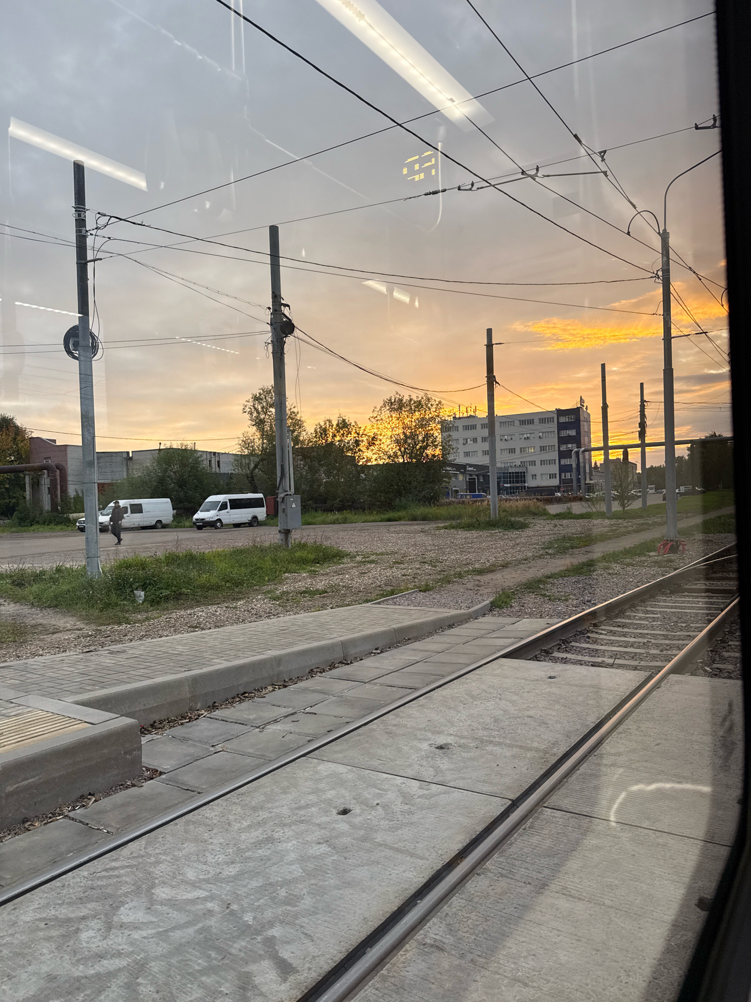The image captures a view from a moving vehicle, likely a tram, looking out towards an urban landscape at dusk. The sky is filled with warm, orange and yellow hues, indicating sunset or sunrise. In the foreground, a tram track runs parallel to the viewer's perspective, with concrete platforms and ballast visible.
The middle ground reveals several objects and structures. Two white minibuses are parked near a low, brick building. A single person, wearing dark clothing, is walking in the vicinity of the minibuses. Further back, a multi-story building with a light-colored facade stands prominently, with trees and vegetation surrounding it. Utility poles with overhead wires crisscross the scene, common in urban areas.
The background is dominated by the sky and some distant trees. The image suggests an industrial or semi-industrial area within a city. Based on the distinctive yellow sign with Cyrillic lettering that reads "Ярославль" (Yaroslavl) visible on the overhead wires in a cropped section of the image, this scene is located in the city of Yaroslavl, Russia. The overall impression is one of a transient urban moment captured during the golden hour.