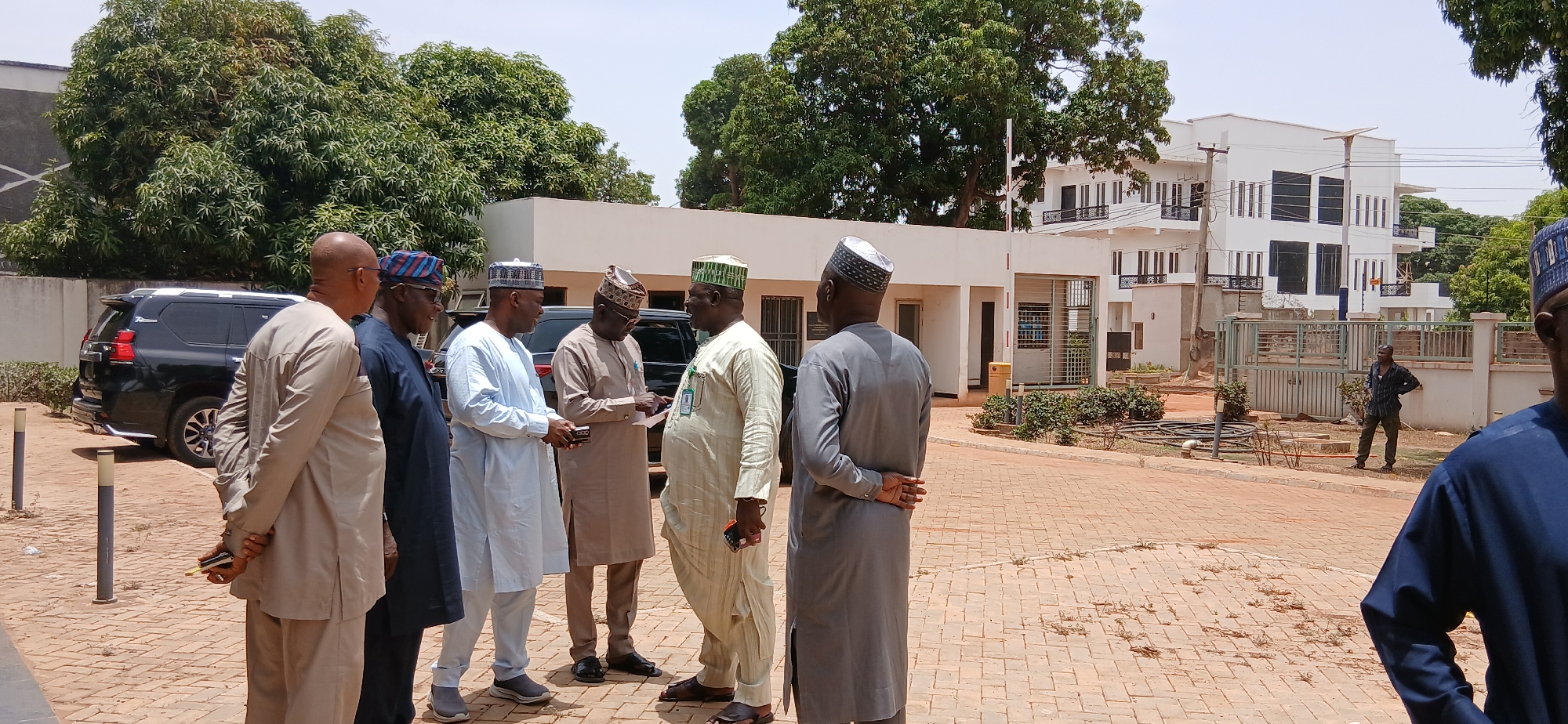 The image shows a daylight scene in Unguwar Shanu, Nigeria, featuring a group of approximately seven men engaged in an outdoor discussion. Most men are dressed in traditional Nigerian attire, comprising long tunics (such as kaftans or agbada) and distinctive caps.
On the far left, a man in a light brown tunic stands with his hands clasped behind his back, holding a mobile phone. Beside him, another man wears a patterned dark blue top and a striped cap. Three central figures appear to be in active conversation; one in a light blue tunic holds a phone, another in a brown tunic examines what appears to be papers or a document, and a third in a light green tunic, wearing a green and white cap, has an ID badge attached to his garment. To their right, a man in a grey tunic stands with his back to the viewer, hands clasped, while another man in a dark blue tunic is partially visible on the far right.
In the background, a black Toyota Land Cruiser Prado SUV is parked. The setting includes lush green trees and modern, multi-story white buildings with balconies, suggesting an office or residential complex. Under a bright, clear sky, a man in a plaid shirt and trousers is seen standing near a gate in the mid-distance. The ground is paved with reddish-brown interlocking bricks. The time of day is clearly bright and sunny, indicating fair weather. The only visible text is the "PRADO" emblem on the rear of the SUV. The overall scene depicts a calm, informal gathering or meeting.