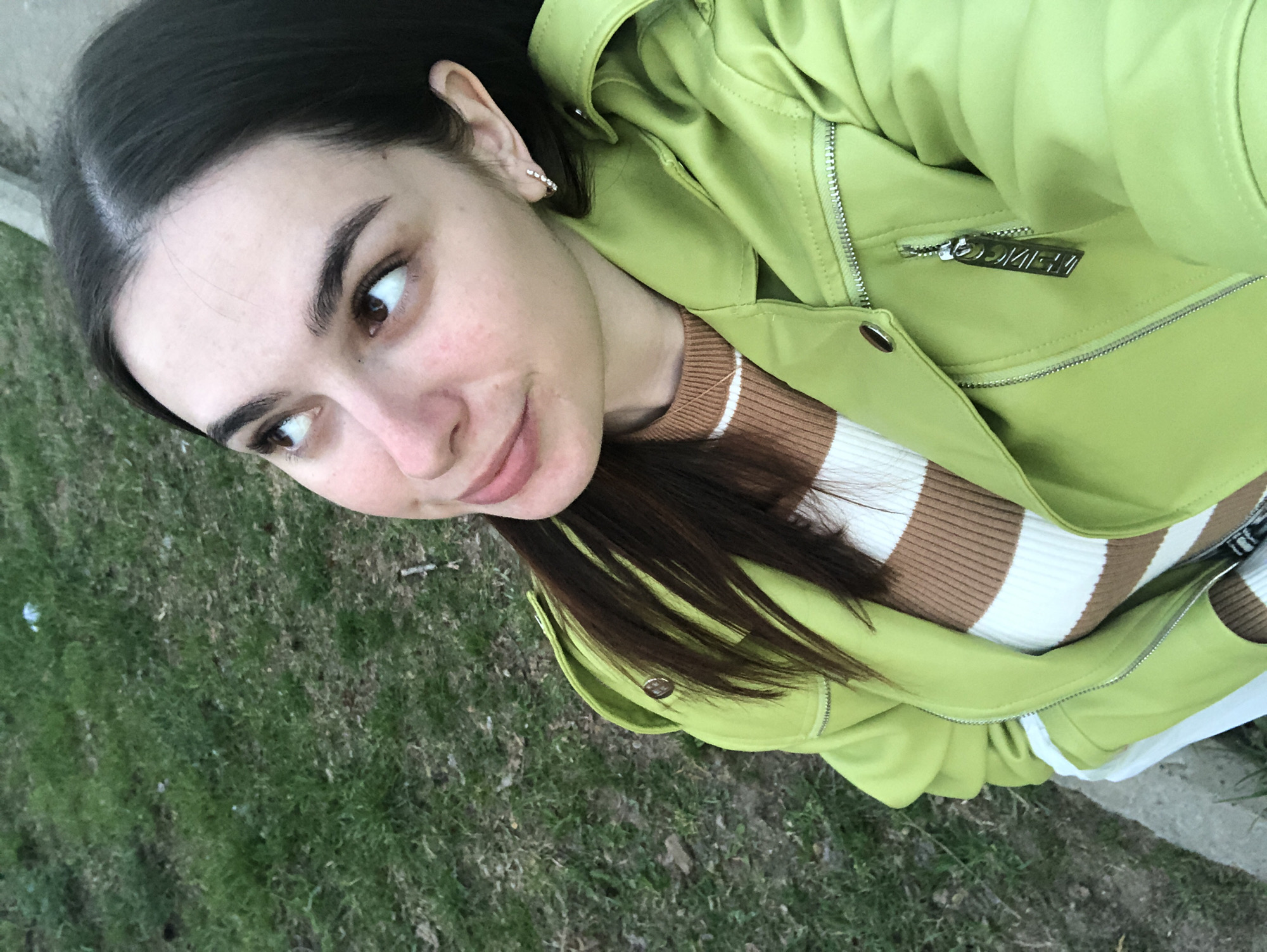 A young woman poses for a selfie outdoors. She has long, dark hair, brown eyes, and fair skin. She wears a brown and white striped sweater under a light green jacket. The jacket appears to be a lighter material and has a zipper and button details. She wears small, possibly diamond, earrings. The background is an area with green grass and some concrete. It is impossible to verify the location.