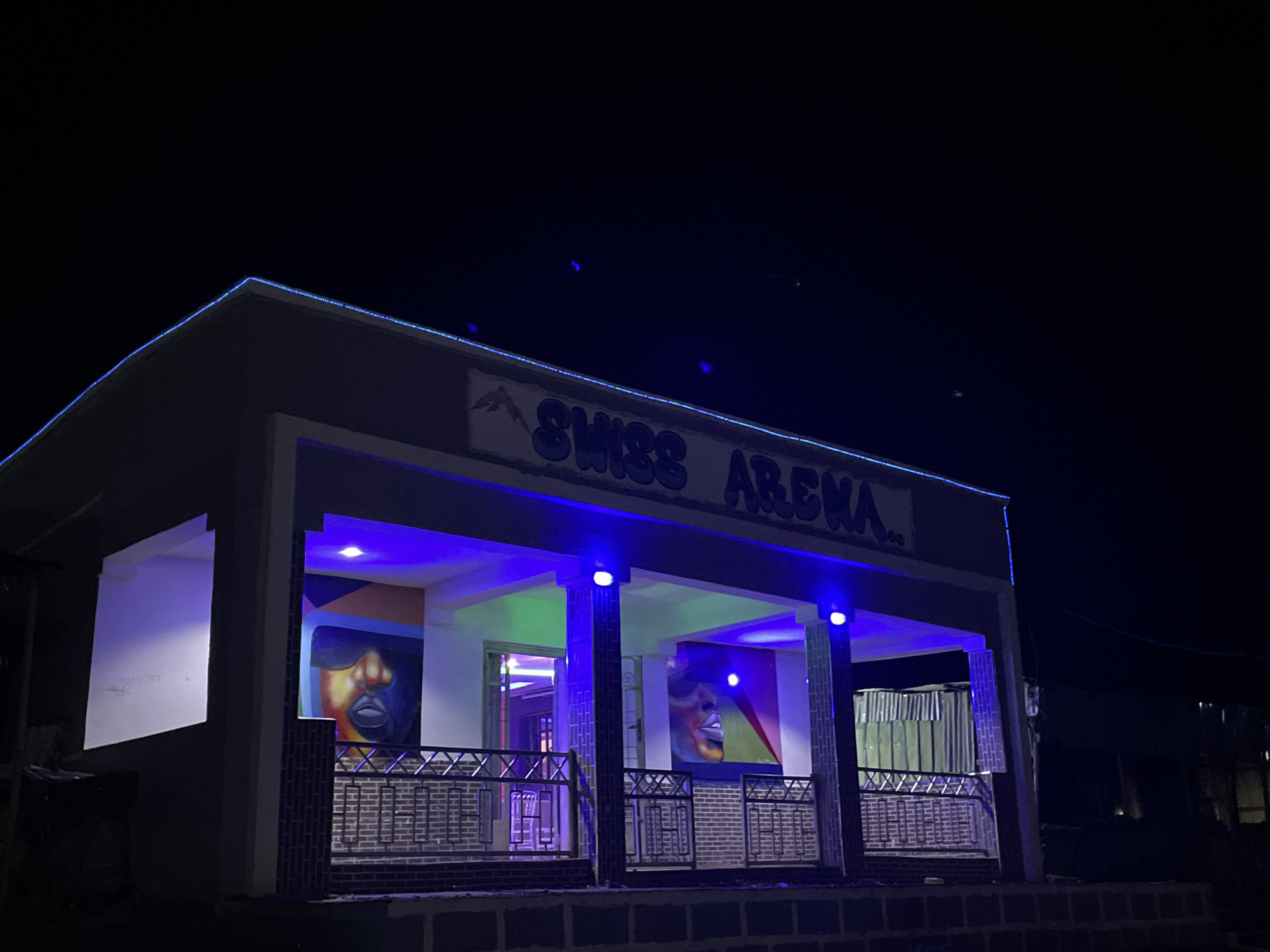 A night-time exterior shot of a building with blue and purple lighting. The building has a sign that reads "SWISS ARENA". There are murals of faces on the walls. The building appears to be in Durumi, Nigeria.