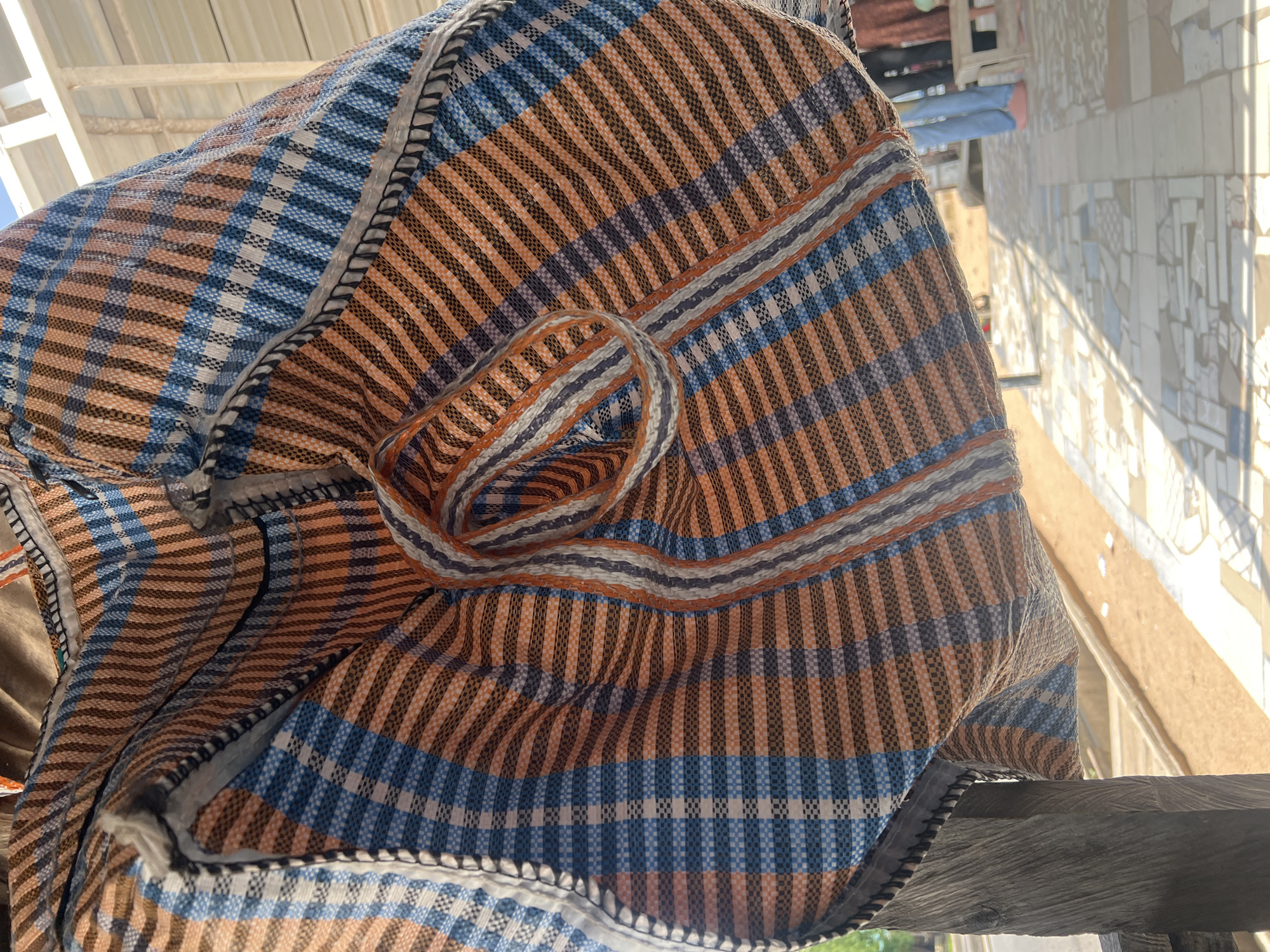 A close-up, eye-level, vertical shot shows a colorful, woven plastic bag hanging from a wooden post in Jalingo, Nigeria. The bag features a plaid pattern of orange, blue, and white stripes. A braided handle made of the same material is visible, coiled and attached to the bag's opening. The background is blurred, suggesting an outdoor environment with sunlight creating dappled shadows on a tiled floor. In the distance, parts of two individuals wearing blue trousers and pink footwear are visible, along with a wooden stool. The scene appears to be in a covered outdoor space, possibly a market or a shaded patio, given the visible corrugated metal roofing overhead.