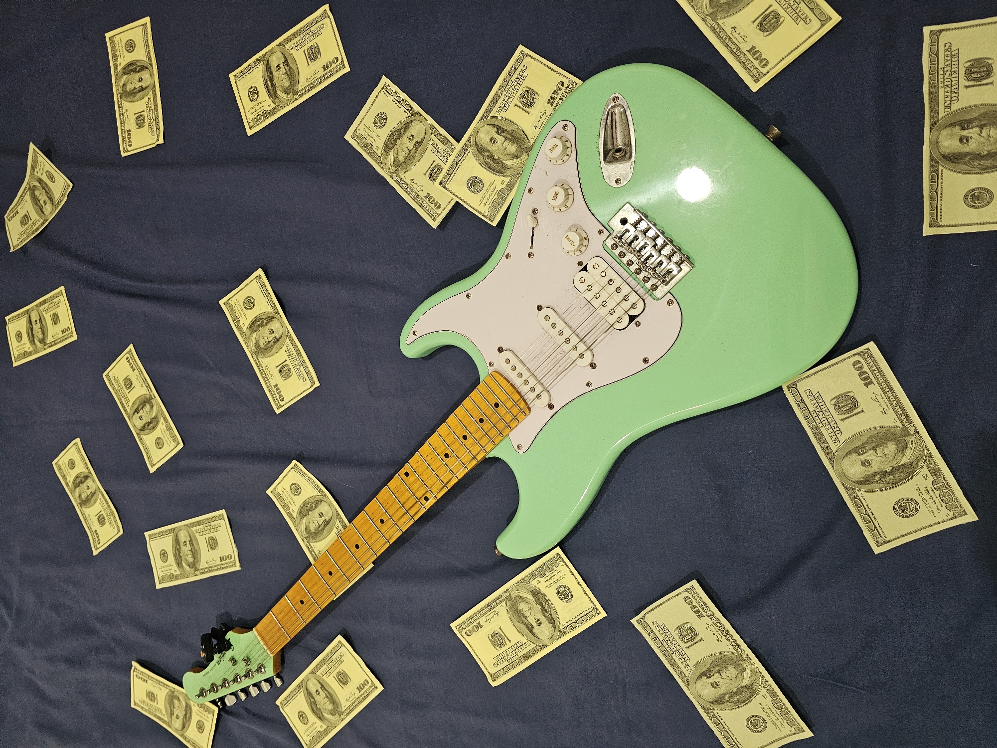 A high-angle, full shot presents a light green electric guitar situated on a dark blue fabric background. The guitar is surrounded by scattered US hundred-dollar bills. The guitar’s headstock is at the lower left corner, while the body is positioned towards the upper right. The overall composition of the scene is disorganized. No details suggest a specific location in Denpasar, Indonesia.