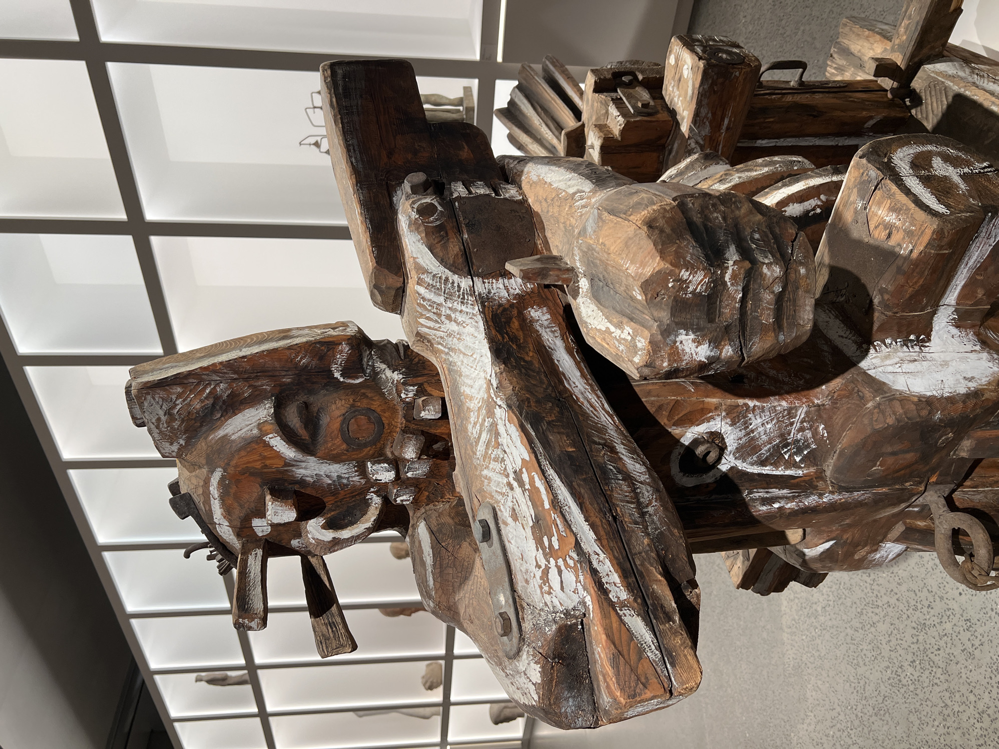 This image captures a large, complex wooden sculpture displayed indoors, likely in an exhibition space or museum in Moscow, Russia. The artwork is composed of numerous roughly carved and assembled pieces of dark brown wood, many of which are accented with strokes of white paint, creating a textured, weathered, and somewhat distressed appearance.
The sculpture features abstract and organic forms; one section resembles a stylized face or head with a circular carving that could be an eye, while another part suggests a clenched fist. Various metal components, such as bolts, plates, and rings, are integrated into the wooden structure, suggesting mechanical connections or reinforcement. The scale of the sculpture appears significant, given its prominence in the frame.
The background consists of a brightly lit, grid-patterned false ceiling with square translucent panels, suggesting diffused artificial lighting. White walls are visible in the distance, further indicating an indoor gallery or contemporary exhibition setting. The perspective is angled, looking up at the sculpture from below and to the side, highlighting its three-dimensional nature and details. No specific activity is depicted, as it's a static display of an art object. There is no visible text in the image.