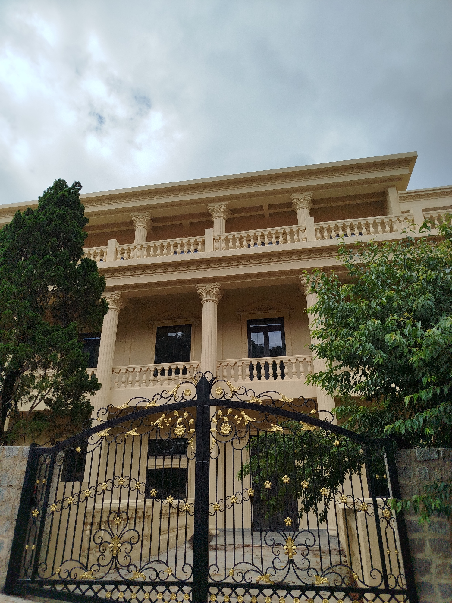 The image depicts a grand, classical-style building with beige walls and columns, likely located in Lạc Dương, Vietnam. In the foreground, a black wrought-iron gate with intricate gold floral embellishments stands closed, obscuring the entrance to the property. Two large trees, one on the left and another on the right, flank the building. The tree on the left is a tall, dark green conifer, while the tree on the right is a deciduous tree with lighter green foliage. The sky above is overcast with scattered white clouds. The architecture suggests a high-end residence or a public building, possibly a hotel or a government office, given the ornate gate and the building's style. The overall scene conveys a sense of opulence and historical influence, characteristic of certain architectural trends found in Vietnam.