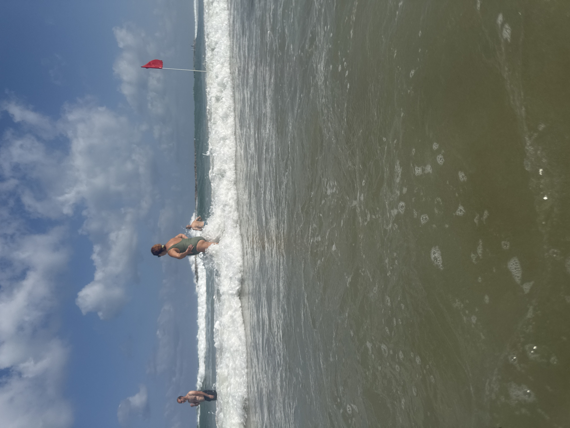A group of people are enjoying the ocean at Herzliya Beach in Herzliya, Israel, under a partly cloudy blue sky. In the foreground, the murky green ocean water is choppy with small waves and foam. In the middle ground, a woman in a green swimsuit is wading in the ocean, with a wave breaking around her waist. To her left, a man with his back to the camera is standing in the water, looking out at the sea. To her right and slightly behind her, another person is partially visible in the water. Further out to sea, a red flag is planted on a white pole, signifying conditions at the beach. The horizon is dotted with distant figures and structures, suggesting other beachgoers or facilities. The overall scene depicts a casual day at the beach with moderate wave action.