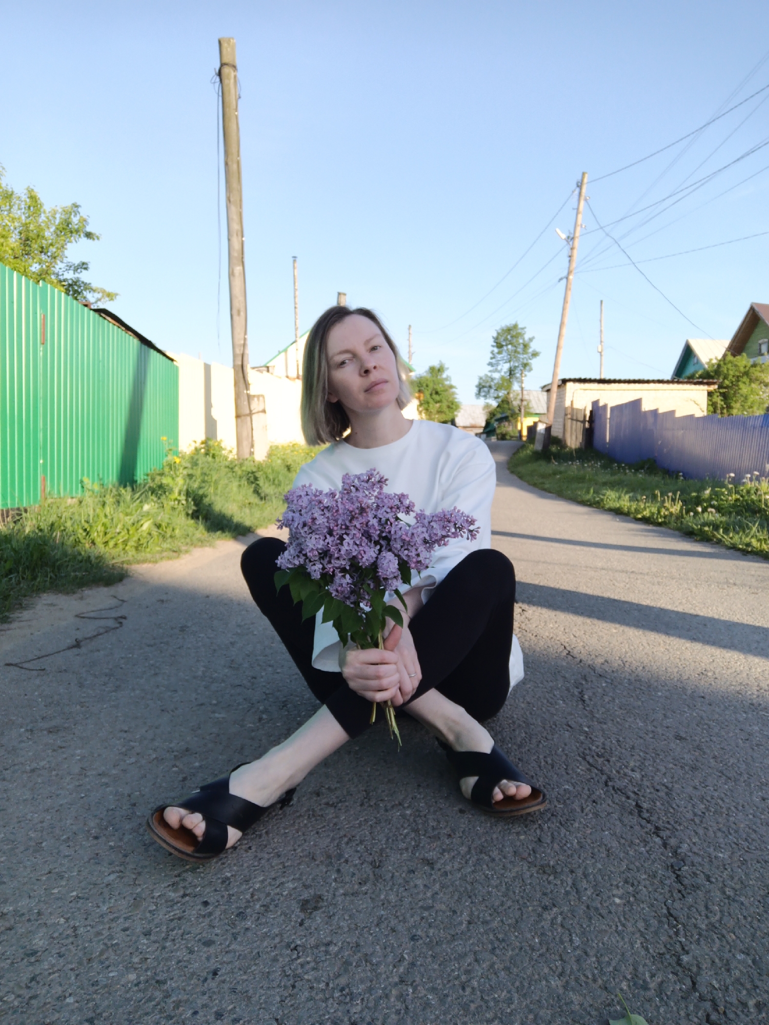 A woman sits cross-legged on an asphalt road in Chaikovsky, Russia. She wears a white sweatshirt, black leggings, and black sandals. She holds a bouquet of purple lilacs. Power lines and wooden poles line the road. Fences and houses are visible on either side of the road. The sky is blue. Green vegetation grows beside a green fence. The lighting is bright, suggesting it is daytime.