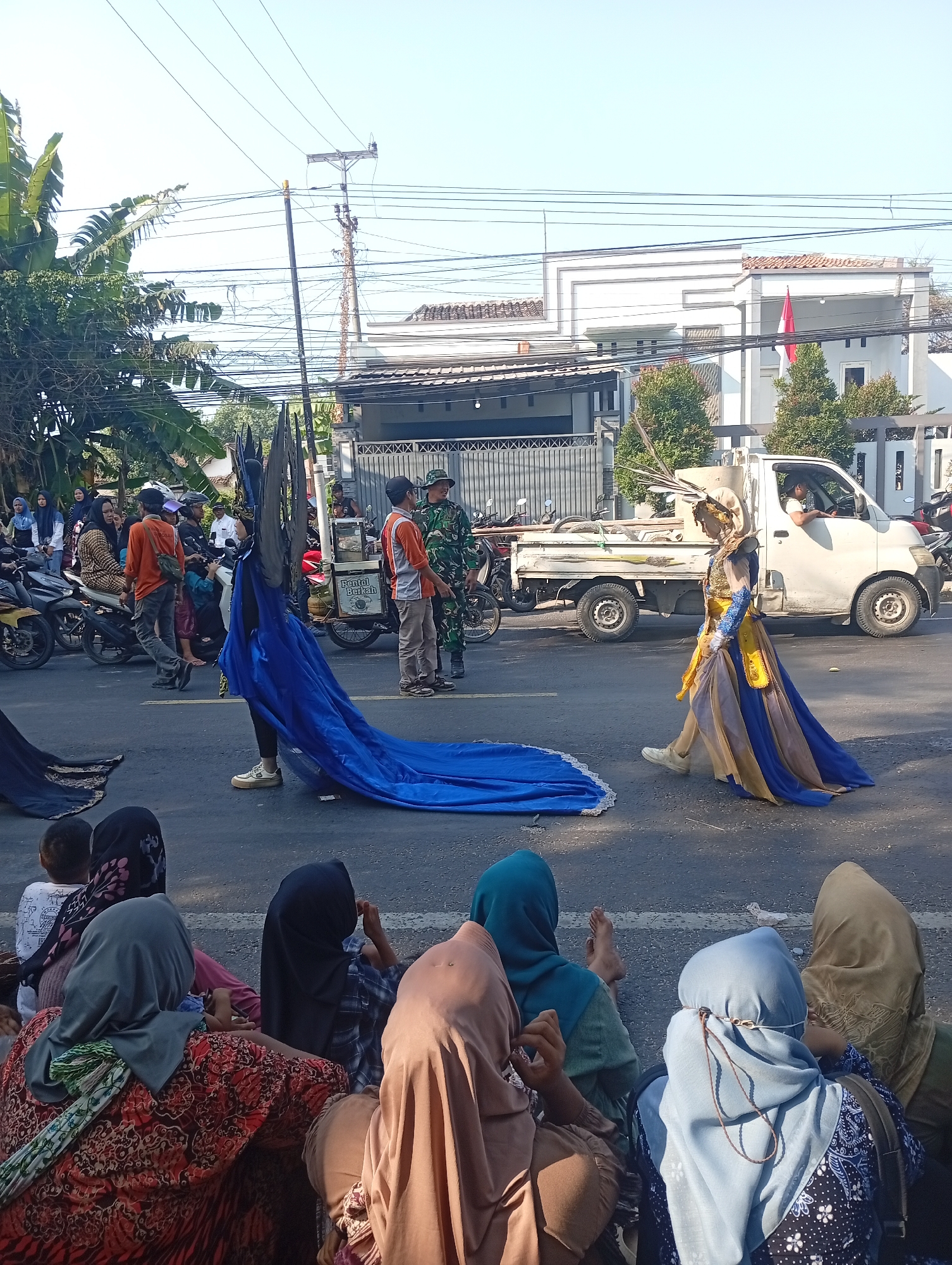The image depicts a street scene in Bajingjowo, Indonesia, where a parade or cultural event is underway. In the foreground, several individuals, primarily women wearing hijabs, are seated on the asphalt, observing the procession. Behind them, the street is occupied by participants and vehicles. A person in an elaborate blue and gold costume with a long train walks across the street. To their left, another person holds a large blue fabric, possibly part of the same costume or a prop.
Further down the street, a white utility truck is visible, with a person driving and another standing nearby. Several motorcycles are parked along the side of the road, and some people are standing around them. A man in a military uniform is also present, interacting with civilians. In the background, houses with tiled roofs and fences line the street, with power lines crisscrossing overhead. The overall atmosphere suggests a public gathering taking place on a sunny day.