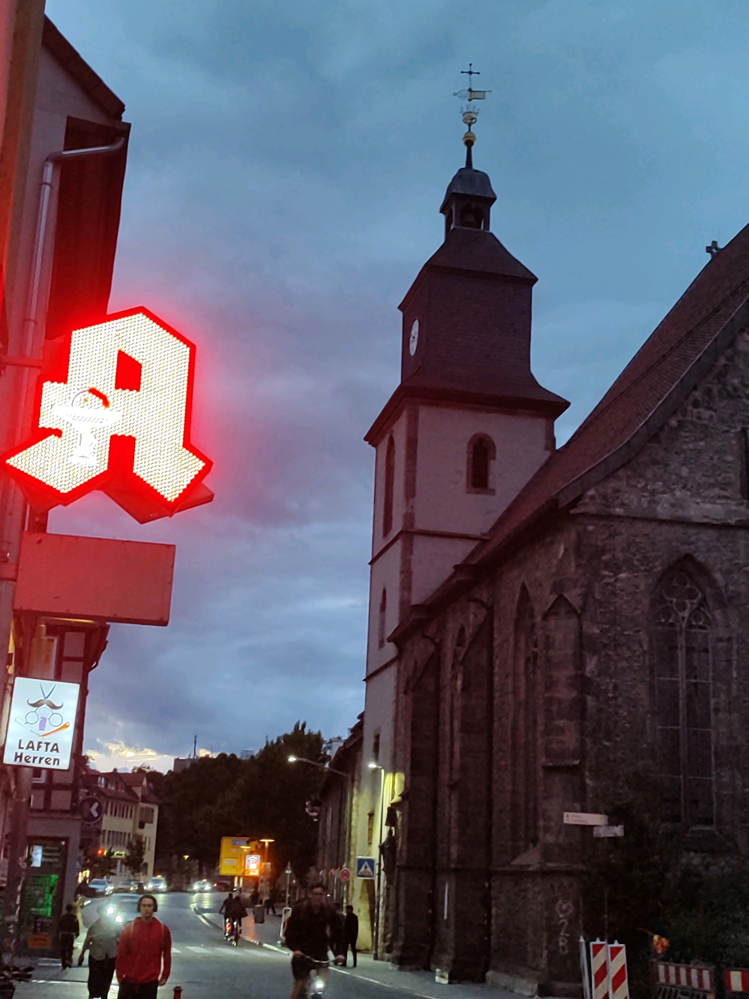 The image depicts a street scene in Göttingen, Germany, during twilight. A prominent red neon "A" sign, likely for an pharmacy, illuminates the left side of the frame. Below it, a smaller sign displays a mustache, scissors, and the text "LAFTA Herren". To the right, a stone church with a steeple dominates the skyline, and a weather vane in the shape of a ship is visible atop the steeple.
The street is moderately busy, with several individuals walking and cycling. One person in the foreground wears a red hoodie and headphones. Cars are present on the street, and streetlights cast a warm glow. The sky is overcast with a mix of grey and dark clouds, suggesting impending weather. The overall atmosphere is urban and slightly moody due to the low light conditions.