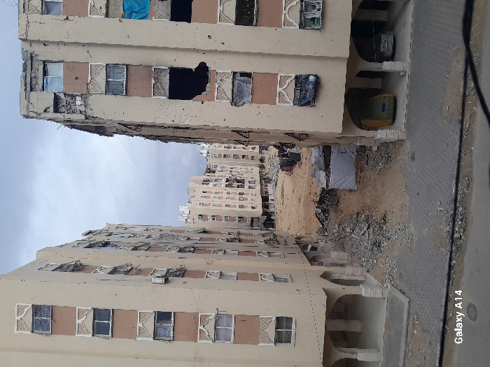 The image depicts a damaged urban residential environment in Khan Yunis, Palestinian Territory, under an overcast sky. Two multi-story residential buildings, primarily light beige with brown architectural accents and decorative window frames, frame a central area of ground-level destruction. The building on the left exhibits multiple broken or boarded windows and scattered facade damage. The building on the right shows more significant structural compromise, with large sections of its exterior facade missing, exposing dark interior spaces; some windows are covered with blue material. At the ground level of the right building, two vehicles are parked in arched recesses: one is covered with a yellow/tan tarp, and the other is a silver sedan. The central ground area between the buildings is an unpaved expanse of light brown dirt heavily strewn with concrete rubble and other debris. A small, temporary structure covered with a light-colored tarpaulin is located towards the center-right of this lot, with clothes hanging on a line or object further back. Additional damaged multi-story residential buildings are visible in the background. No individuals are present.
