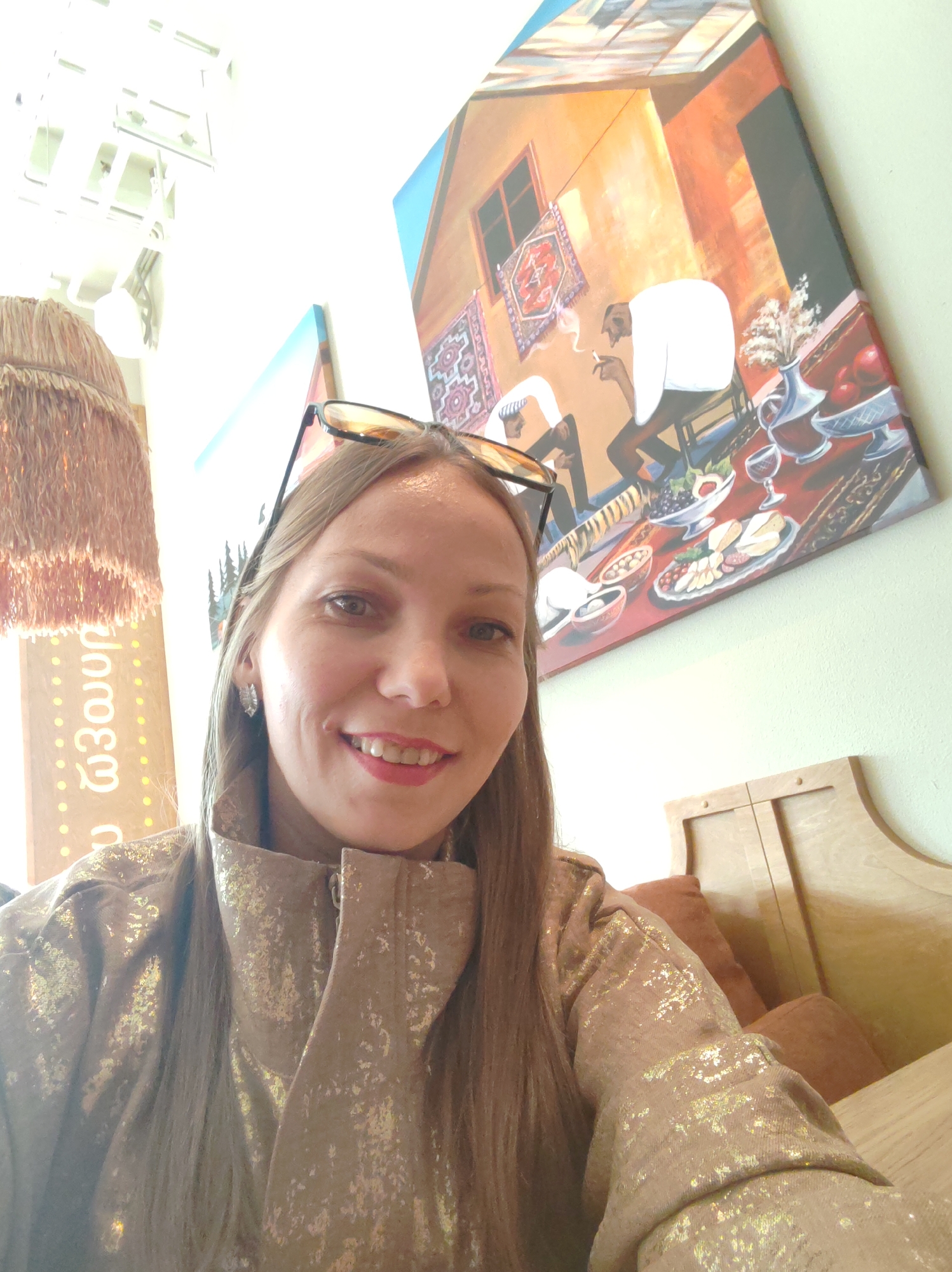 A blonde woman with long, straight hair and sunglasses perched on her head smiles at the camera. She is wearing a shiny, gold-flecked jacket. Behind her, a colorful painting depicts two figures, one smoking, seated at a table laden with food and drinks. To the left of the painting, a wooden sign with yellow dots and Georgian writing is visible, next to a woven lampshade. The background appears to be a light-colored wall, with a portion of a wooden booth on the right. The scene is set indoors, likely in a restaurant or cafe. There is no discernible information in the image to suggest a specific location in Syktyvkar, Russia.