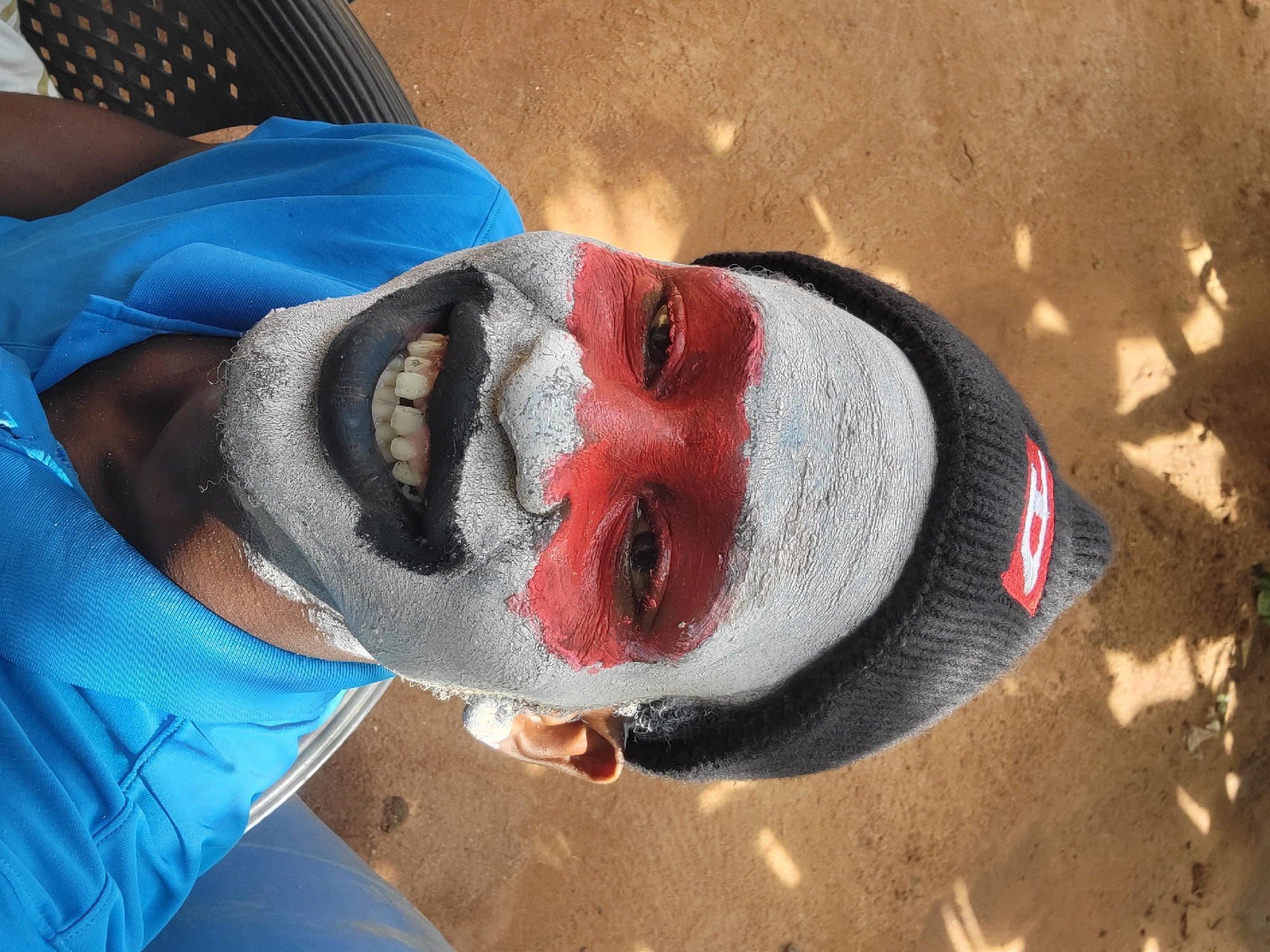 An overhead shot shows a person in the city of Nkpor, Nigeria, wearing a black beanie and a blue polo shirt. Their face is painted white with red paint around their eyes and black paint around their mouth. The person is seated in a black plastic chair. The ground is reddish-brown earth and has some shadows.