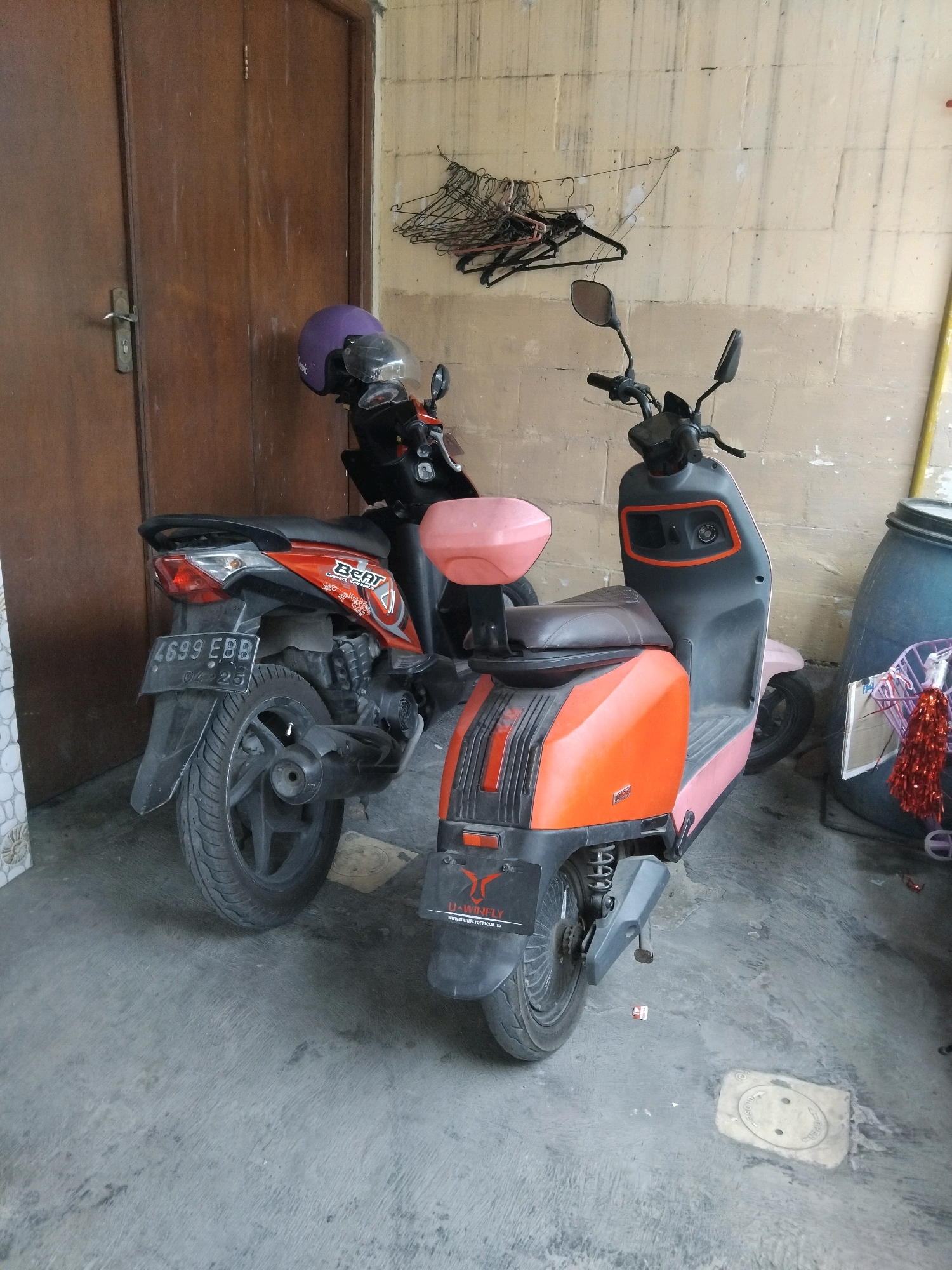 The image depicts a side view of two parked scooters in a covered outdoor area, likely a garage or carport, next to a weathered, light-colored block wall. A wooden door is visible on the left. The scooter in the foreground is predominantly orange and pink with black accents, featuring a pink seat and a small pink storage box on the back. It has a black rear fender with a sticker displaying a red winged logo and "U-WINFLY" text with a URL. A portion of its front wheel and suspension are visible. The scooter behind it is orange and black, with a black seat and a purple helmet resting on its handlebars. This scooter has a license plate reading "4699 EBB" and appears to be a Honda Beat based on the "BEAT" branding on its side. Both scooters are parked on a concrete floor that shows some staining and debris. To the right, a large dark blue barrel is partially visible, along with a yellow pole extending vertically. A cluster of black wire coat hangers is mounted on the wall above and behind the scooters.