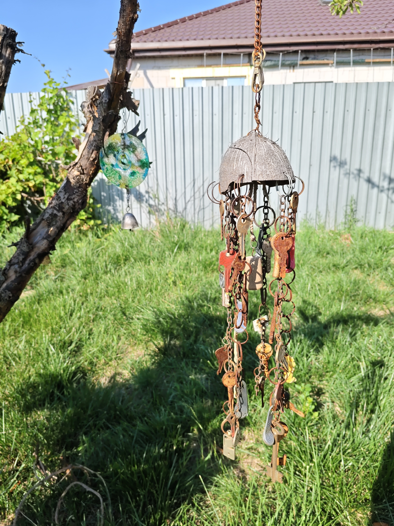 The image depicts a garden scene, likely in Rostov-on-Don, Russia, on a clear day. The foreground is dominated by a tree branch on the left, from which a decorative hanging object is suspended. This object appears to be a wind chime made from a coconut shell, adorned with numerous old and rusty keys attached by chains. To the left and slightly higher on the tree branch, another circular decorative item with blue and green hues is hanging, along with a small silver bell.
The background consists of a green lawn with shadows cast by sunlight. Behind the lawn, a grey corrugated metal fence runs horizontally across the image. Above the fence, a section of a building with a brown tiled roof is visible, with a light-colored wall and a window. The sky above is a clear, bright blue. There are no people or animals visible in the scene, and the focus is on the decorative items and the natural environment.