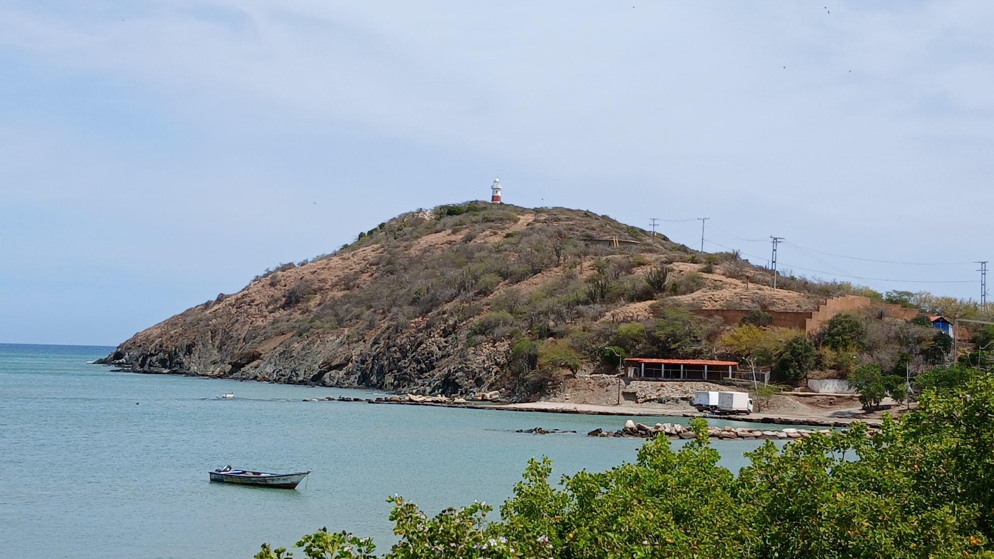 The image presents a panoramic view of a coastal area in Pedro González, Venezuela, under a clear, light blue sky. A large, arid hill dominates the central and right portions of the frame, characterized by sparse, brownish scrub vegetation, cacti, and exposed rock formations. A red and white striped lighthouse with a white lantern room is prominently situated atop this hill. Multiple utility poles with visible power lines traverse the upper right slope of the hill.
The hill's base meets a tranquil body of light blue-green water, identified as a bay or cove, which extends to the left and into the background. A rocky coastline delineates the transition from the hill to the water. In the foreground water, slightly left of center, a single small, open fishing boat with a visible outboard motor is anchored; it displays white, blue, and red hull markings. Another smaller, white object floats further out in the water to the left.
On the immediate shoreline, at the base of the hill on the right, a single-story building with a reddish-brown tiled roof and open sides is present. Adjacent to this structure, two white box trucks are parked on an unpaved, sandy area. Further up the hillside, behind these vehicles, a large, earth-toned terraced structure is integrated into the slope, with a smaller building featuring a blue roof visible to its right. The immediate foreground is partially obscured by dense green foliage. No individuals are discernible within the scene.