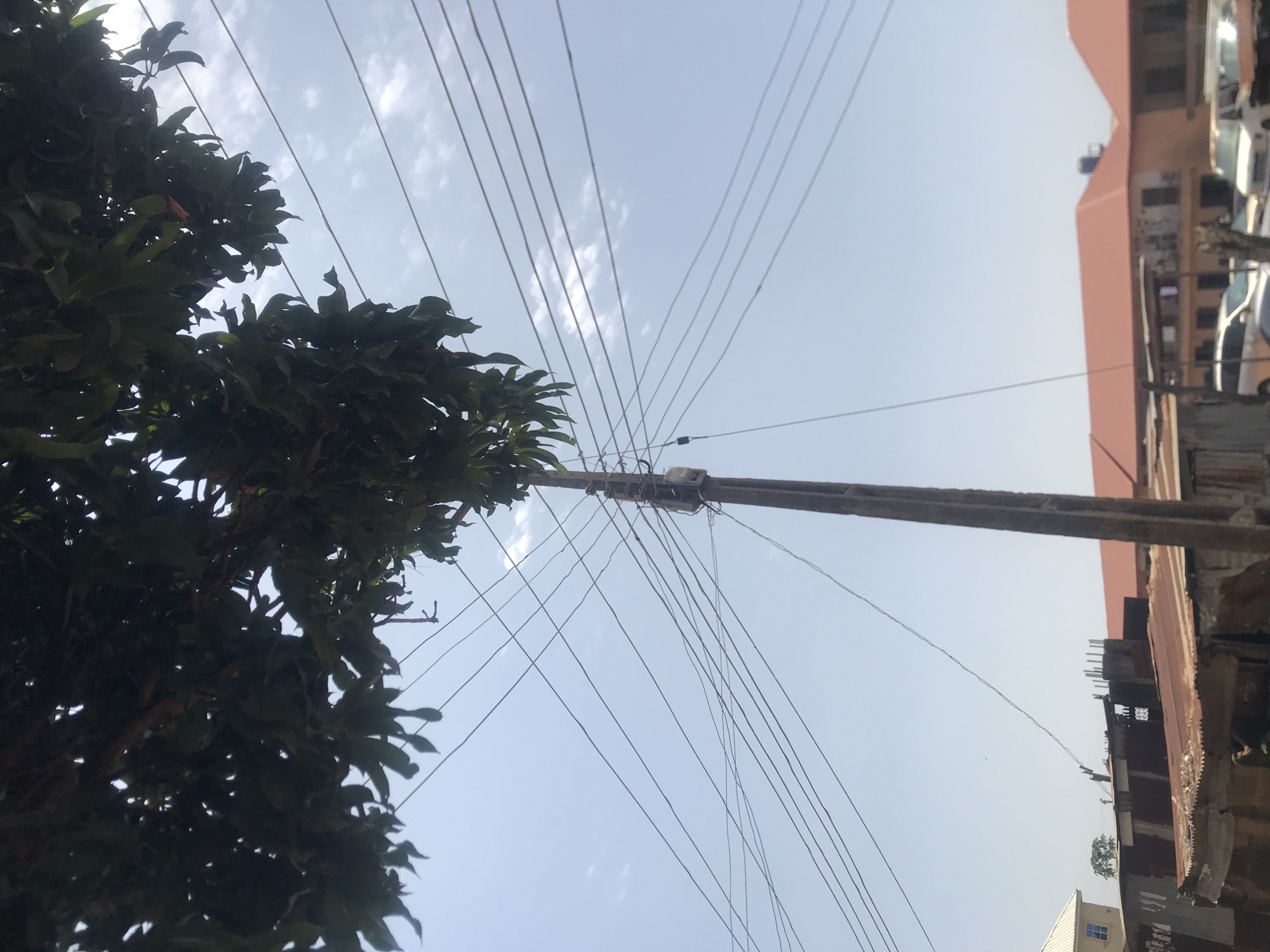 This is an outdoor, eye-level shot featuring a utility pole with multiple overhead wires and a box attached to it. The pole is positioned in the center-right of the frame. Overhead, a dense cluster of dark green leaves from a tree canopy fills the upper portion of the image, obscuring the sky in places. Below the tree, the sky is a pale blue with scattered white clouds. In the background, a row of buildings with corrugated metal roofs and some orange-tiled roofs are visible. Several vehicles, including white vans and what appear to be cars, are parked along the street in front of the buildings. The overall scene suggests a residential or commercial area in a city. The location is identified as Rafin Par, Nigeria.