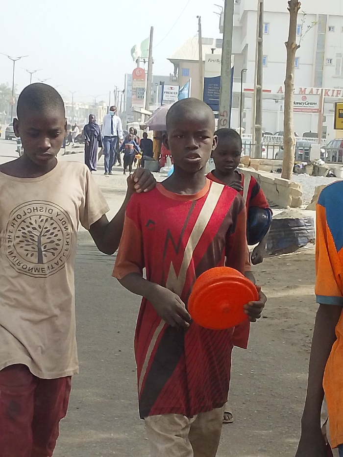 The image depicts an outdoor daytime scene on an urban street in Gandu Albasa, Nigeria. In the immediate foreground, three young males are visible. The leftmost boy, with dark skin and short hair, wears a light brown short-sleeved t-shirt featuring a circular logo with a tree design and mirrored text. He wears dark trousers and looks towards the lower left. The central boy, also with dark skin and short hair, wears a red short-sleeved t-shirt with black, white, and lighter red geometric patterns and light-colored trousers. He holds an orange plastic bowl in his left hand and places his right hand on the left shoulder of the boy to his immediate left. This central boy looks directly towards the viewer. The third boy, partially visible on the right, wears an orange and blue shirt.
In the midground, several pedestrians are present on what appears to be a sidewalk or dirt path adjacent to the street. A man in a blue suit, white shirt, and light blue face mask walks away from the viewer, accompanied by a woman in a dark, flowing garment (abaya/hijab). Other indistinct figures are also walking.
The background reveals a bustling street lined with multi-story buildings. Visible signs include "FirstBank," "PREMIER," "AL MAI KIFIEN," and "MoneyGram." Utility poles, one marked with the number "65," and streetlights are present. Green and white flags, likely Nigerian, are displayed on a pole. Cars are parked or moving in the distance. The atmosphere appears dusty or hazy, suggesting dry weather conditions. Bare tree branches are also visible.