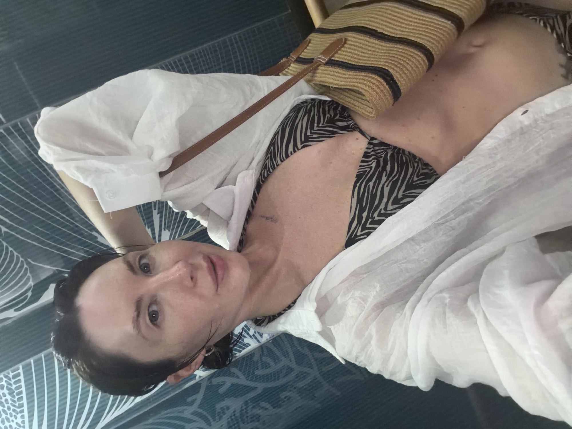 A woman with dark hair and fair skin takes a selfie in what appears to be a restroom with a patterned blue shower curtain. She is wearing a white unbuttoned shirt over a black and white zebra-print bikini top and a woven straw purse with brown straps is slung over her shoulder. The woman is looking directly at the camera with her left arm raised behind her head.