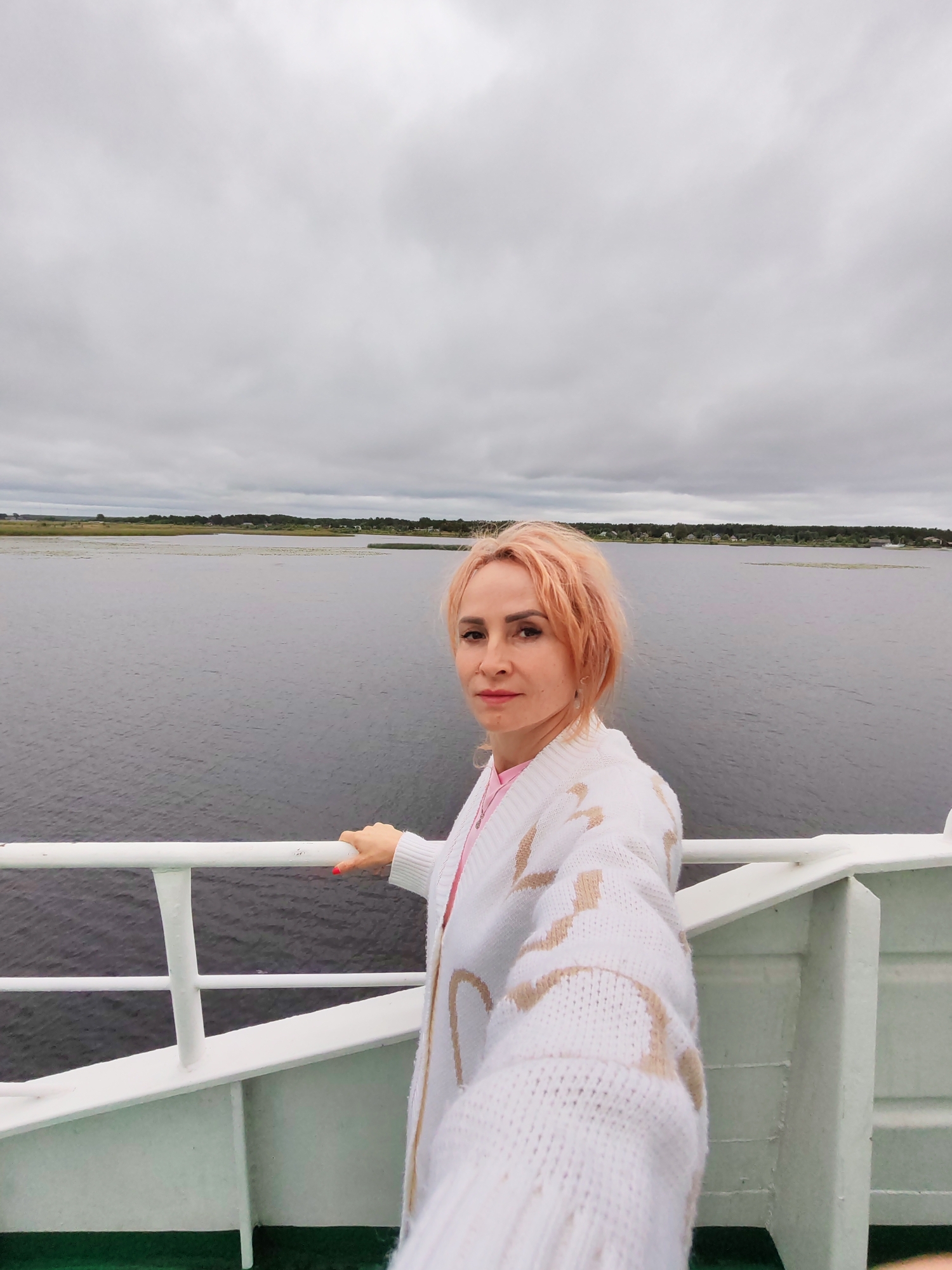 A woman with short, peachy-pink hair and fair skin is taking a selfie on a white ferry. She is wearing a white knit cardigan with a tan pattern over a pink top. Her left hand is resting on a white railing, and her fingernails are painted red. The background is a large body of dark water, with a distant, green, tree-lined shore dotted with small houses. The sky is overcast with gray clouds. This scene is likely on the Neva River or Gulf of Finland, near Shal'skii, Russia, given the landscape and ferry.