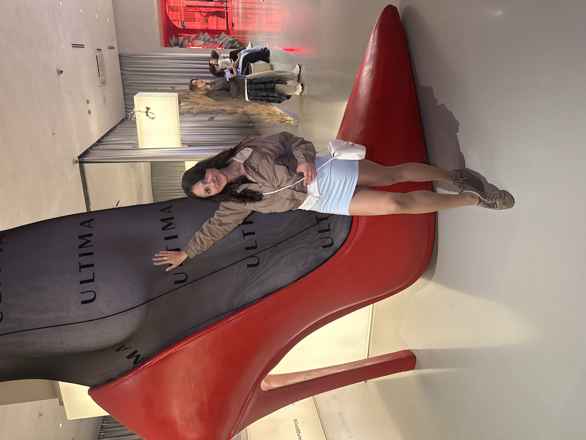 A young woman with dark hair stands leaning against a massive red stiletto heel sculpture in what appears to be a museum or exhibition space in Saint Petersburg, Russia. The sculpture is a dramatic representation of a high-heeled shoe, with a black stocking material forming the leg portion. The woman is wearing a light brown jacket, a pale blue mini-skirt, and beige sneakers. She carries a small white crossbody bag. In the background, other individuals are visible, some seated and others standing, engaged in conversations or looking at displays. The environment features modern architecture with neutral-colored walls, large mirrors, and ambient lighting. The floor is smooth and reflective.