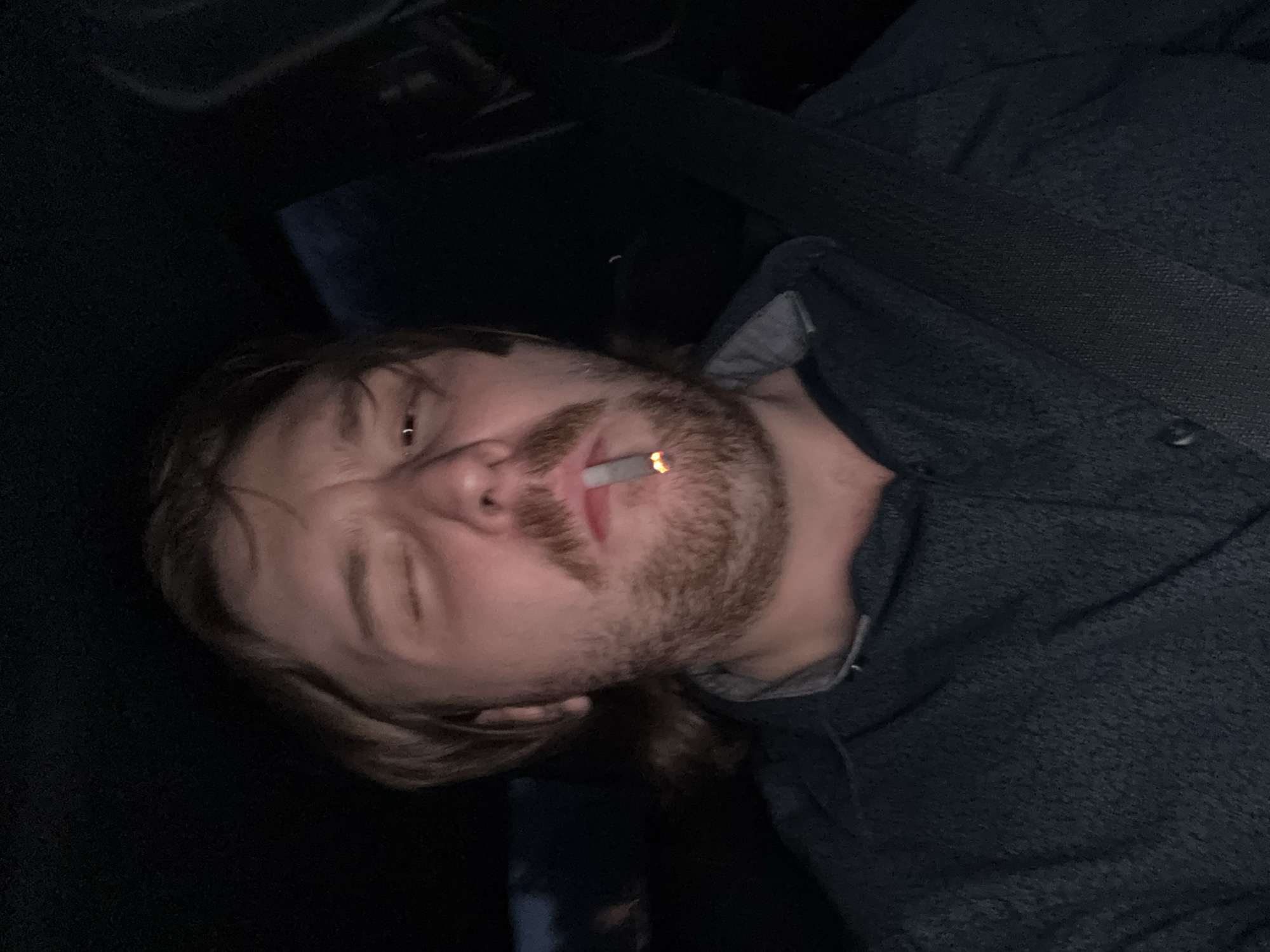 The image shows a man with light brown hair and a beard, wearing a dark blue button-down shirt with a light gray collar. He is positioned at an angle, facing the camera with a cigarette in his mouth. His left eye is squinting. The background is dark, with a portion of what appears to be a seatbelt visible. There is no visible information to determine whether the image was taken in Huixquilucan de Degollado, Mexico.