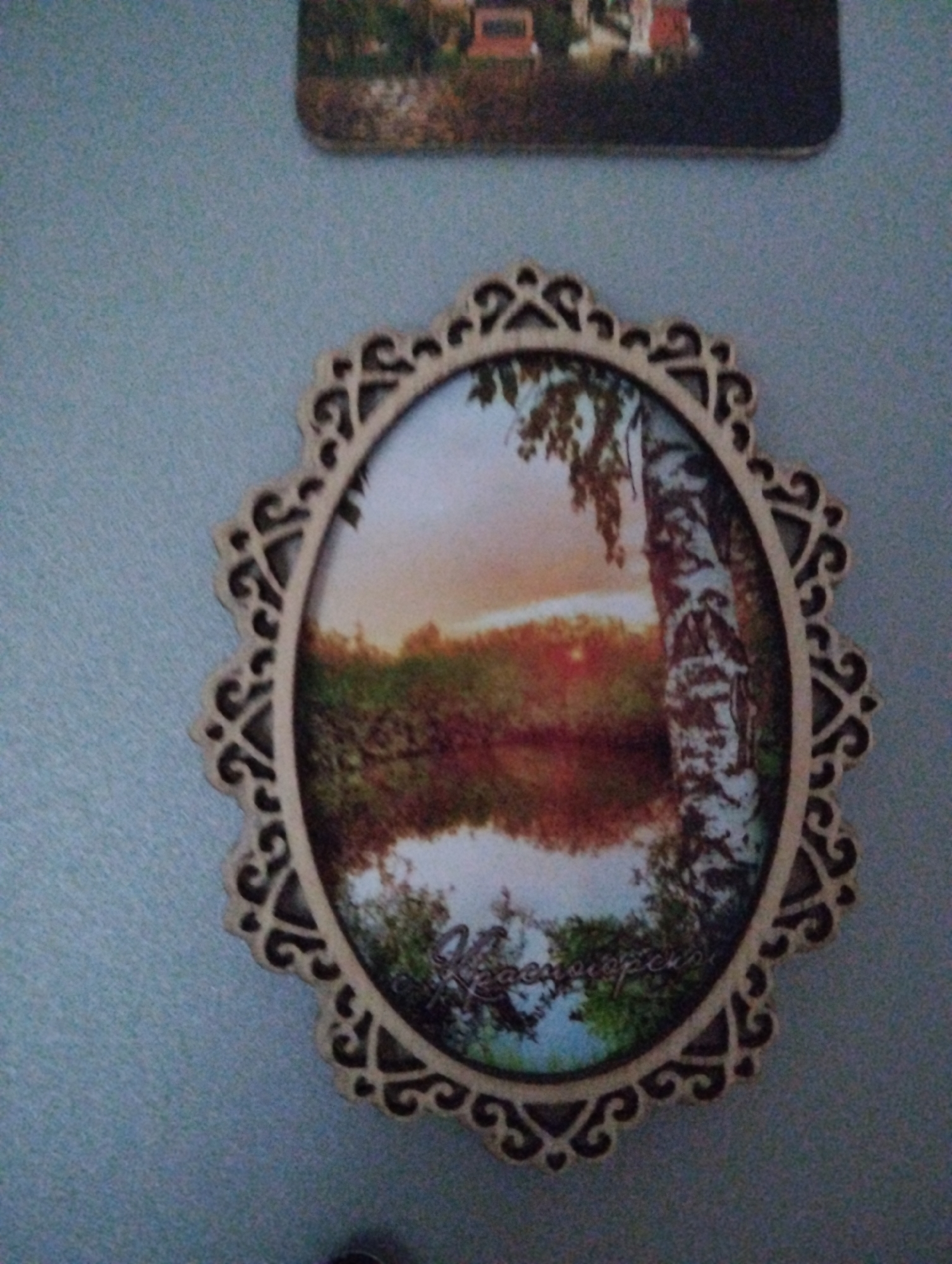 The image shows a framed oval picture of a landscape scene. The scene depicts a lake or pond with trees reflected in the water. A birch tree is visible on the right side of the frame. The sky suggests either a sunrise or sunset. The frame is decorative, with a light brown color and intricate cut-out patterns. The framed picture is mounted on a light blue wall, likely inside a building. There is a partial view of another picture mounted above it. Text can be read on the picture, and it reads "Krasnogorsky." The location is implied, Krasnogorskoye, Russia.