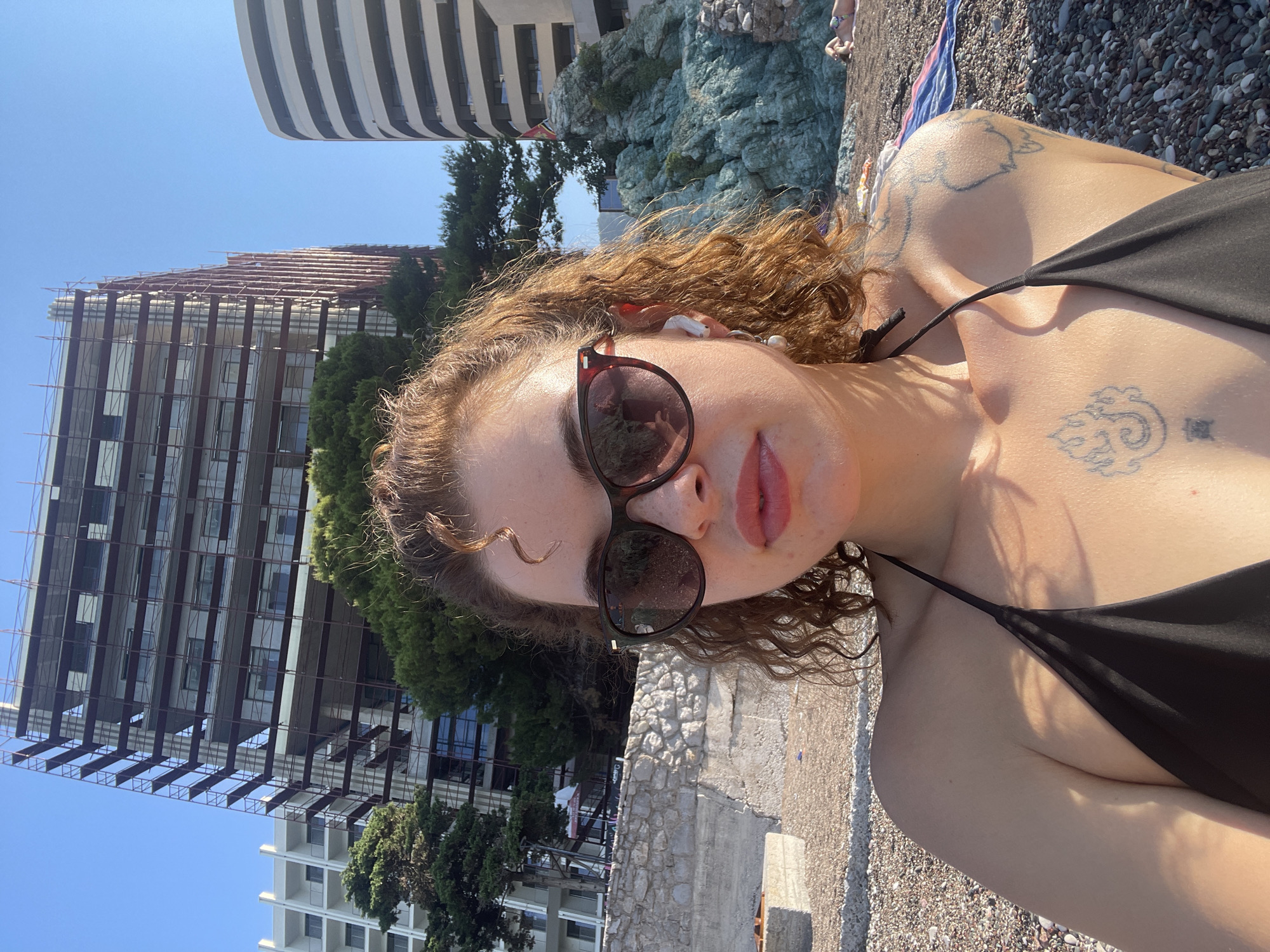 The image depicts a woman with curly, reddish-brown hair, wearing sunglasses and a black bikini top, taking a selfie on a pebble beach. She has a tattoo on her chest and shoulder. In the background, there are modern buildings under construction and a rocky formation with some vegetation. The sky is clear and blue, suggesting a sunny day. The overall setting appears to be a coastal area, potentially in or near Rafailovići, Montenegro, given the presence of multi-story buildings characteristic of coastal resorts and construction activity.