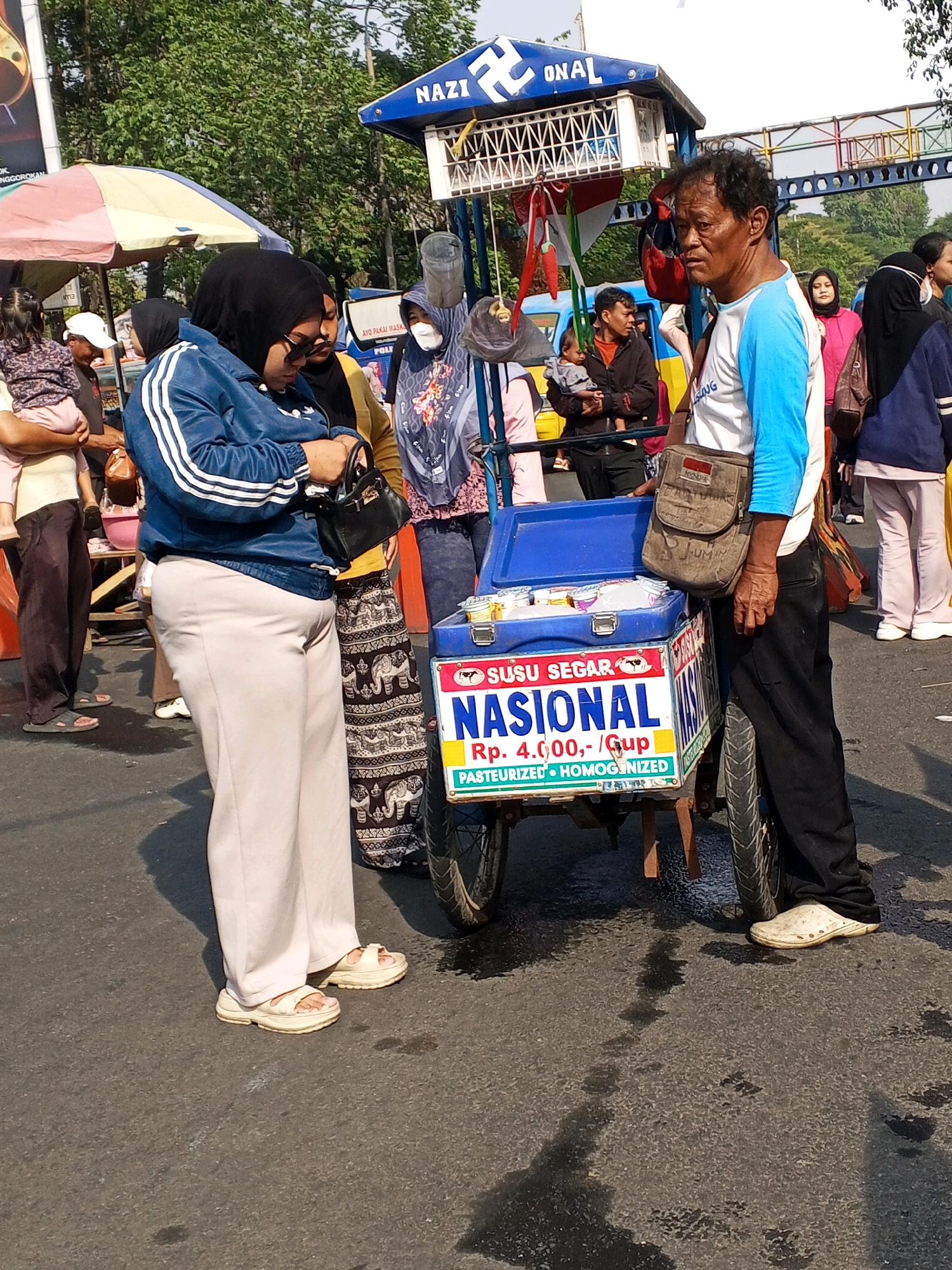 In the city of Serang, Indonesia, a street scene unfolds with a focus on a mobile ice cream cart. The cart itself is adorned with a blue and white sign that reads "NASIONAL" in large blue letters, accompanied by "SUSU SEGAR" (Fresh Milk) at the top, and a price of "Rp. 4.000,-/Gup". Below this, it further specifies "PASTEURIZED • HOMOGENIZED". The cart is equipped with a blue cooler containing several smaller containers of ice cream.
A man, appearing to be the vendor, stands to the right of the cart. He has dark, curly hair and is wearing a blue and white long-sleeved shirt, black pants, and white slip-on shoes. A brown messenger bag is slung across his chest. He is looking off to the side with a neutral expression.
To the left of the cart, two women are engaged in conversation, or perhaps the vendor is speaking to them. The woman in the foreground is wearing a black hijab and sunglasses, a blue jacket with white stripes on the sleeves, and light beige wide-leg pants with beige platform sandals. She is holding a small black handbag. The woman behind her is wearing a hijab and a patterned skirt with elephants and geometric designs, and is holding a dark object.
Other individuals are visible in the background, including children and adults, some partially obscured by the cart and other market stalls. An umbrella with red, yellow, and pink panels is also visible to the left. The ground is asphalt, with some wet patches reflecting the sunlight. Trees line the background, suggesting an outdoor setting, likely a busy street or market area in Serang. The overall atmosphere suggests a typical day in a bustling Indonesian market.