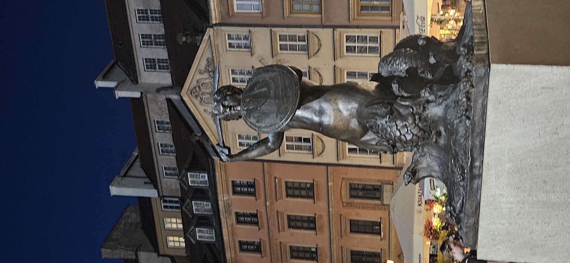 The image captures a bronze statue of the Warsaw Mermaid, a prominent landmark in the city. The statue stands on a stone pedestal in the Old Town Market Square, Warsaw, Poland, against a backdrop of colorful historic buildings and a dark blue night sky. The mermaid is depicted with a sword and shield, a symbol of the city's coat of arms. The sculpture is detailed, showing scales on her lower body and a determined expression on her face. In the foreground, a person is partially visible, leaning towards the base of the statue. The background reveals the facades of old European-style buildings with numerous windows, some illuminated from within, and awnings of shops or cafes. The overall scene suggests a public square at night, with the statue illuminated and the surrounding architecture adding to the historical ambiance.
