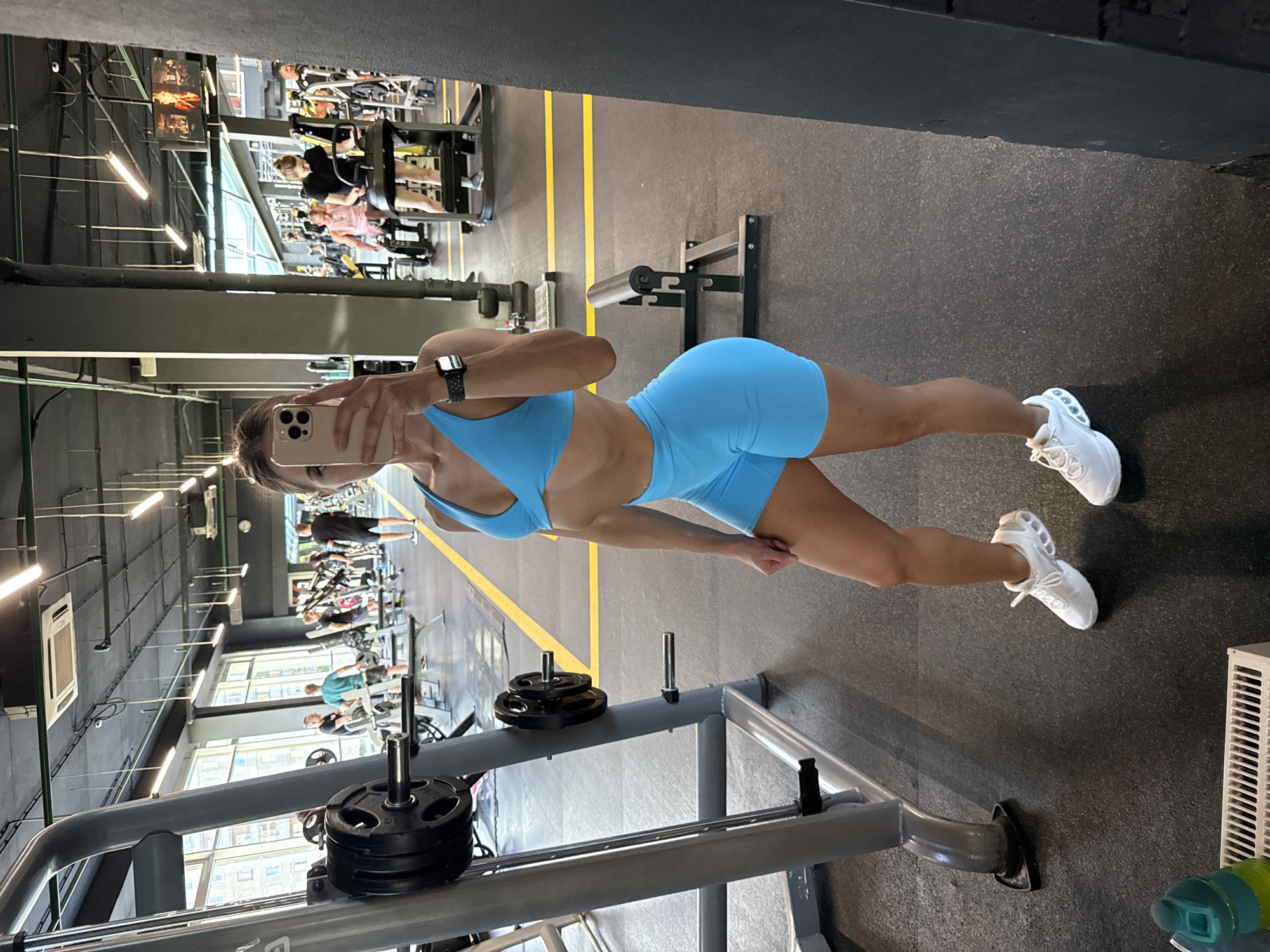 A woman in a blue athletic set and white sneakers is taking a mirror selfie in a gym. She is standing in front of a weight rack. In the background, other gym-goers are visible using exercise equipment. The gym appears to be spacious with large windows. The overall scene is indicative of a fitness center in Saint Petersburg, Russia.