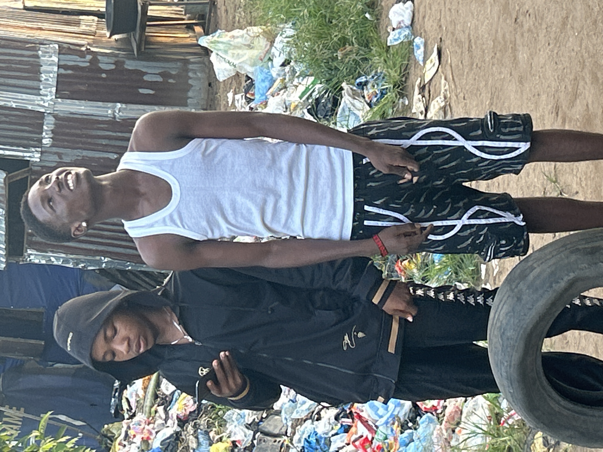 Two young Black males stand in a debris-strewn outdoor area in Ihiagwa, Nigeria. The male on the left, wearing a black hooded jacket and black pants with white detailing, holds a mobile phone and looks down at it. He has a silver chain around his neck. The male on the right, wearing a white tank top and black patterned shorts with white detailing, looks upwards and to the right, with his mouth open. A red bracelet is visible on his left wrist. Behind the individuals, a large pile of trash and discarded items are visible, with some greenery growing in patches. A corrugated metal structure stands in the background, along with a small wooden table and a black container. In the foreground, a large tire is partially visible. The ground is a mix of dirt and sand.